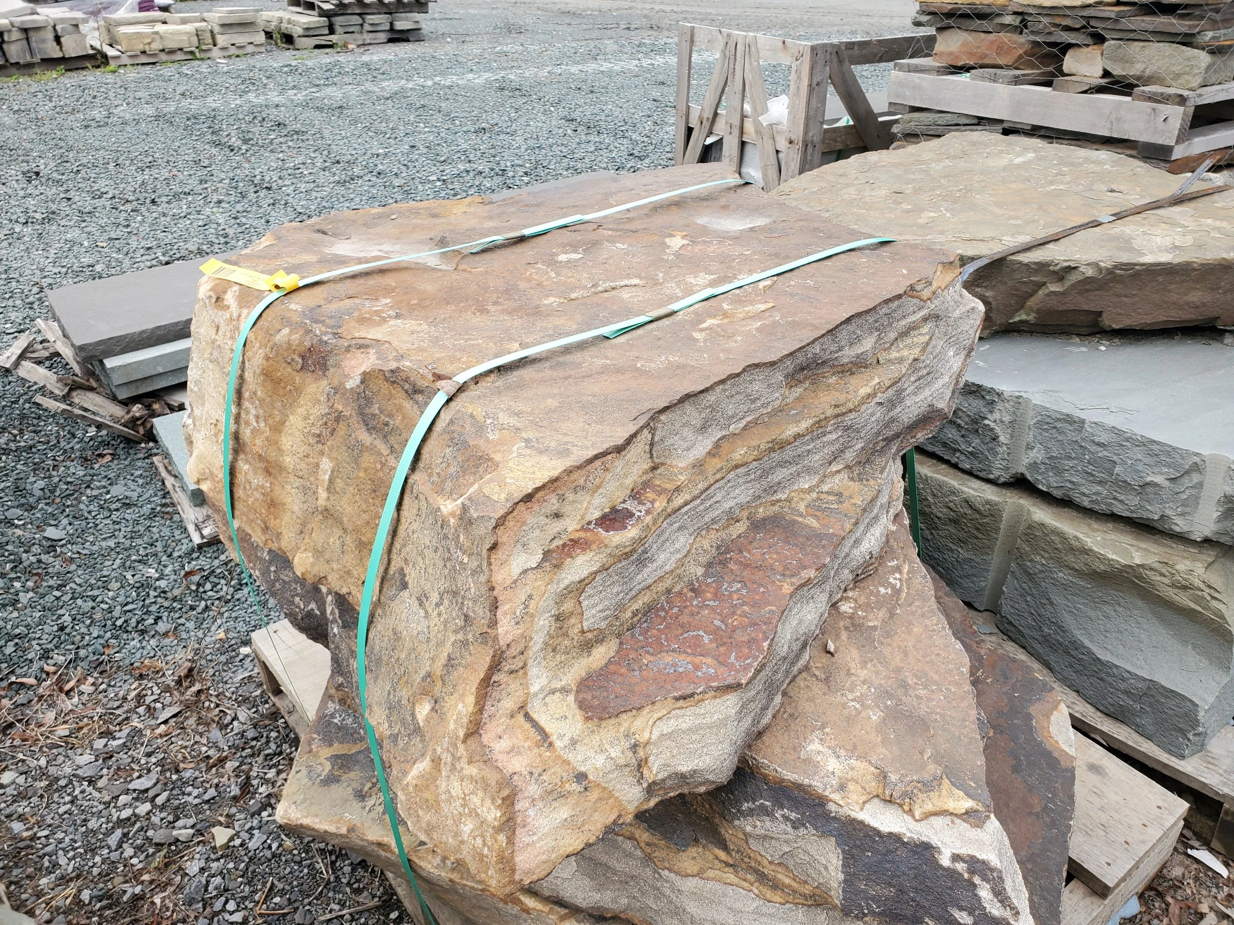 Large natural stone slab on a wooden pallet at an outdoor stone yard, with other slabs and gravel visible in the background.