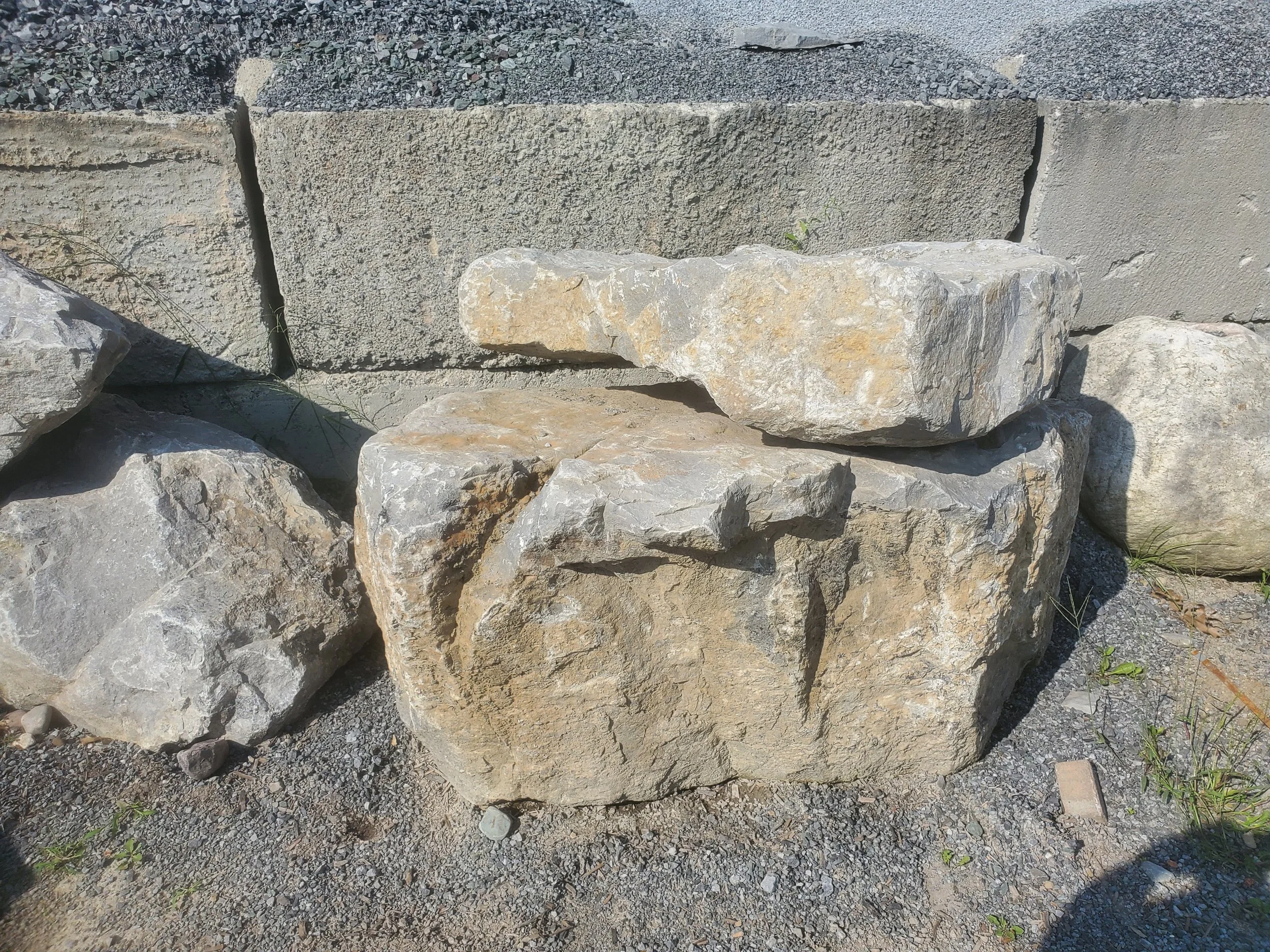 Several large, irregularly shaped rocks and stones placed outdoors against a cement block wall, surrounded by gravel and small plants.