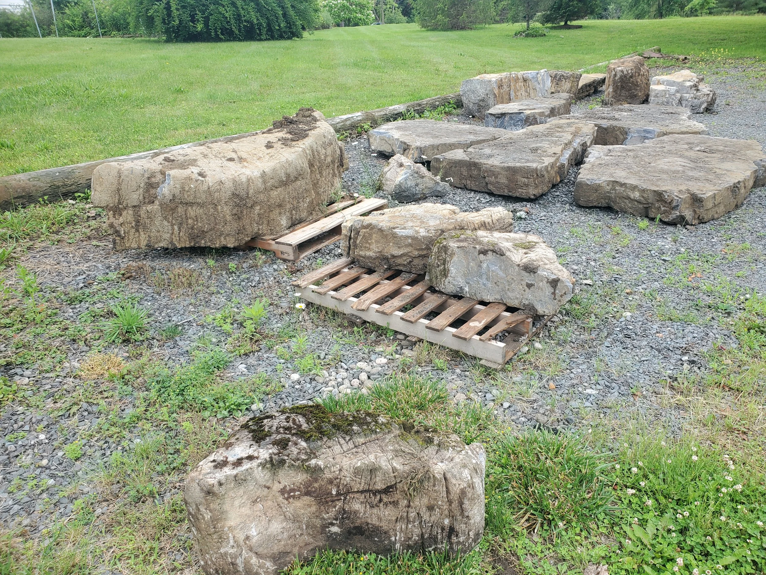 A collection of large rocks and stones placed on a gravel surface with some wooden pallets and a log in a grassy outdoor area.