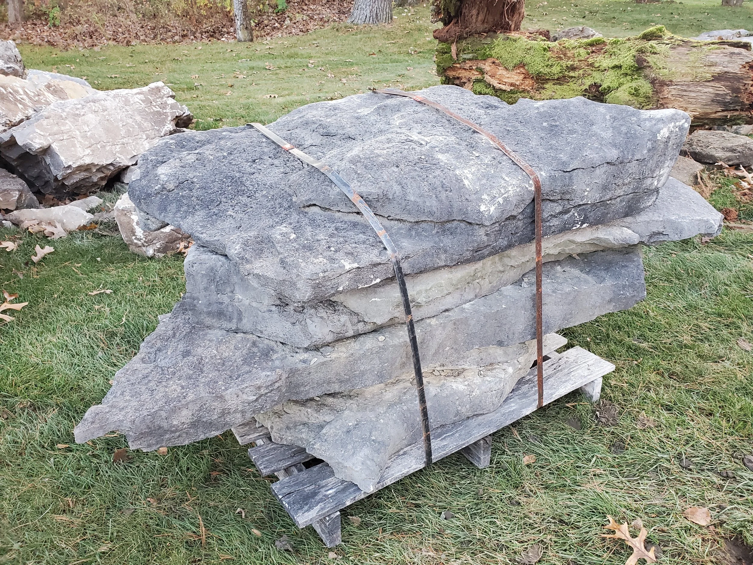 Large, flat rocks stacked on a wooden pallet outdoors, secured with metal straps, with grass and tree trunks in the background.