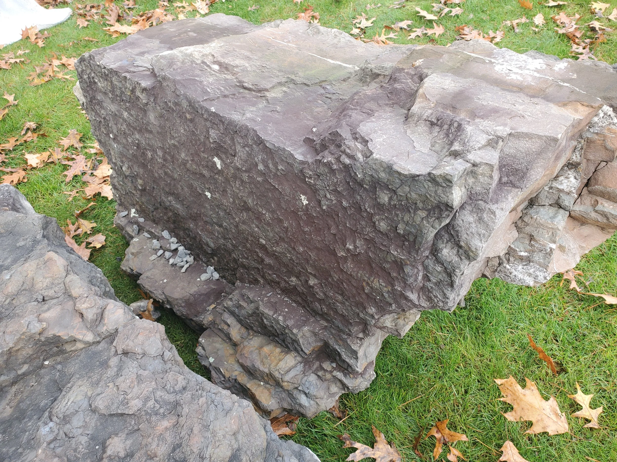 Large, rough-textured gray and brown stone slab placed on grass among fallen autumn leaves.