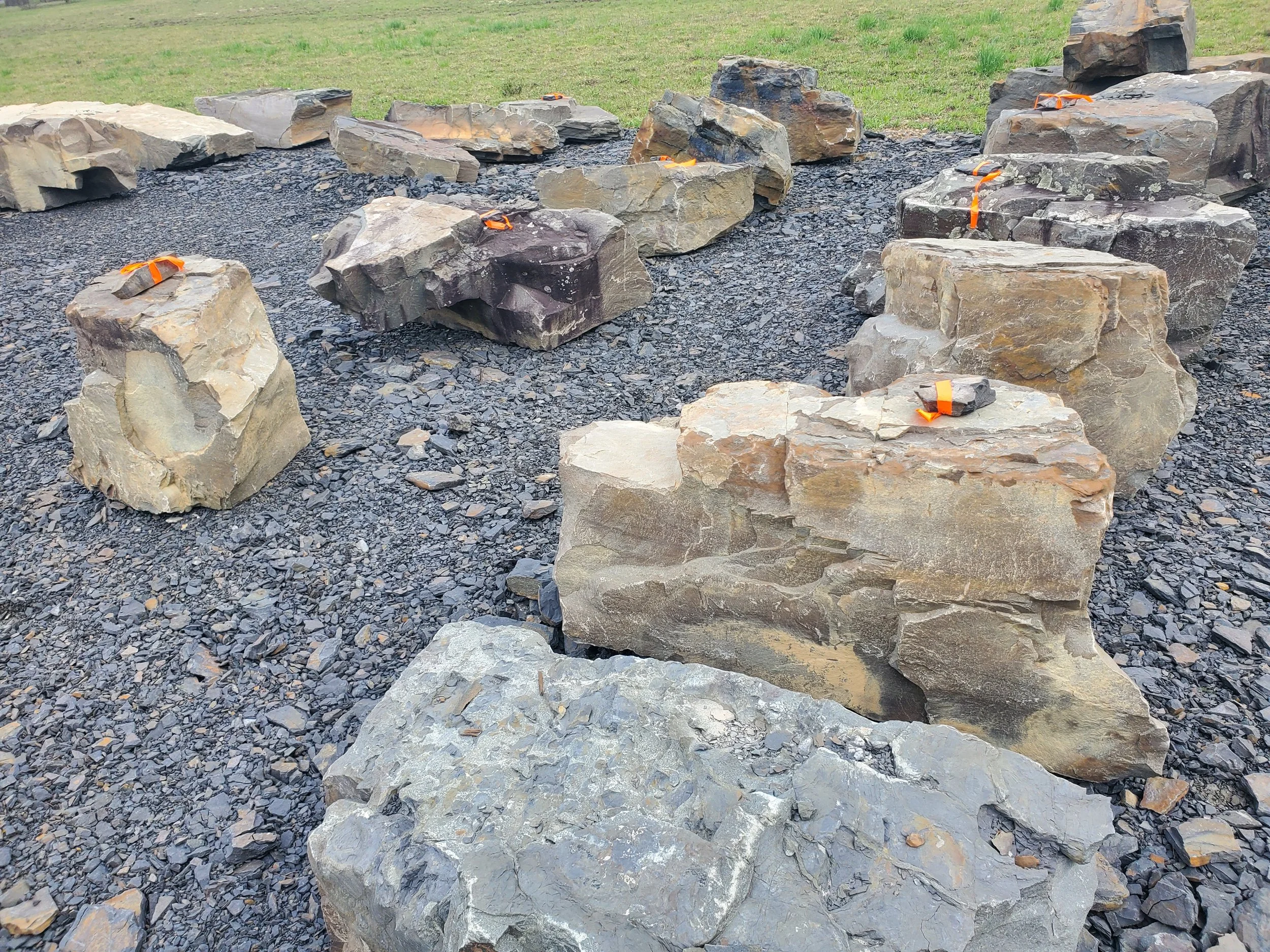 A collection of large, irregularly shaped rocks secured with orange straps, placed on a bed of small black gravel, with a grassy area in the background.