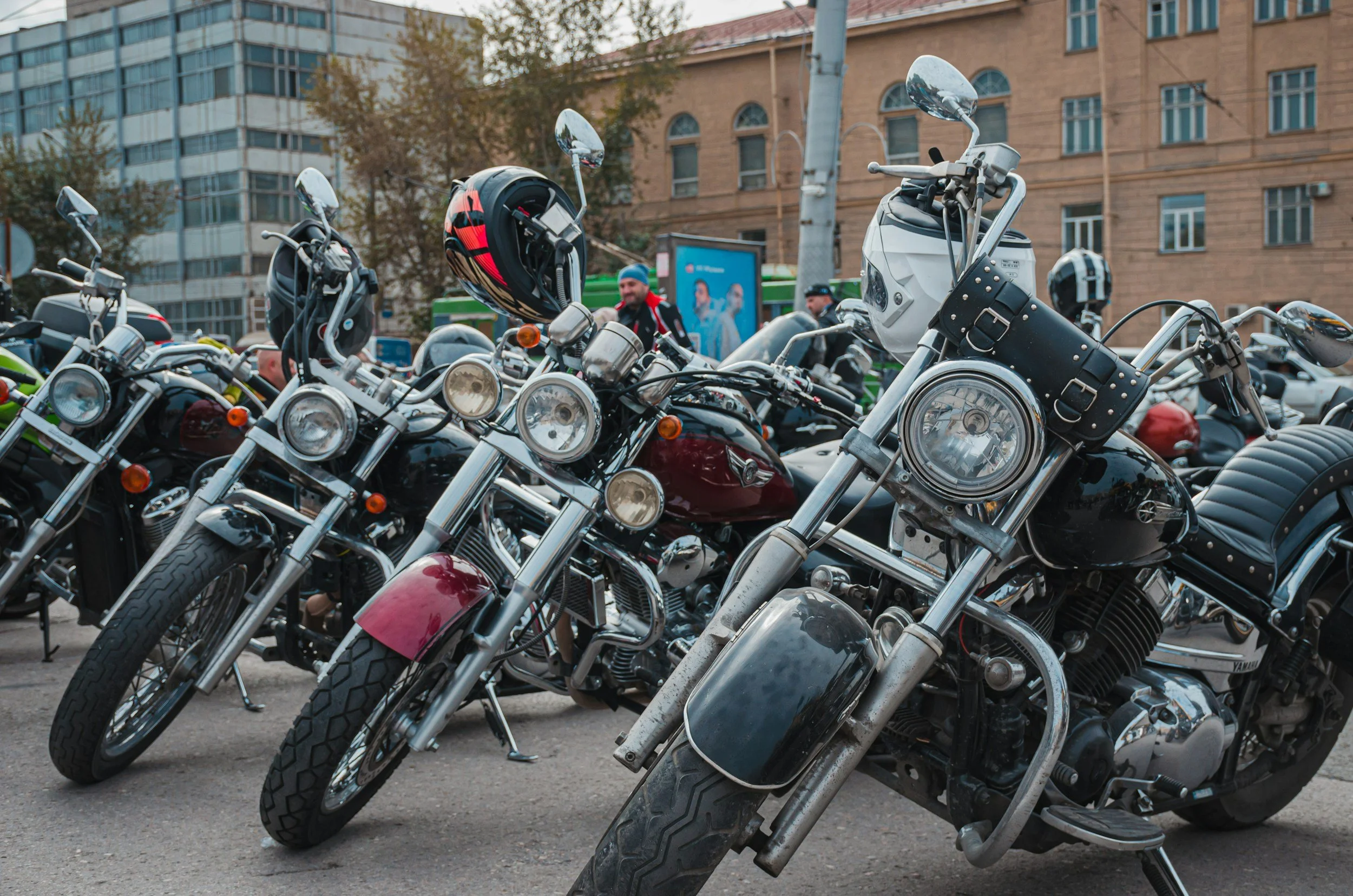 Row of parked motorcycles, some with helmets on handlebars, in an urban setting with buildings and people in the background.