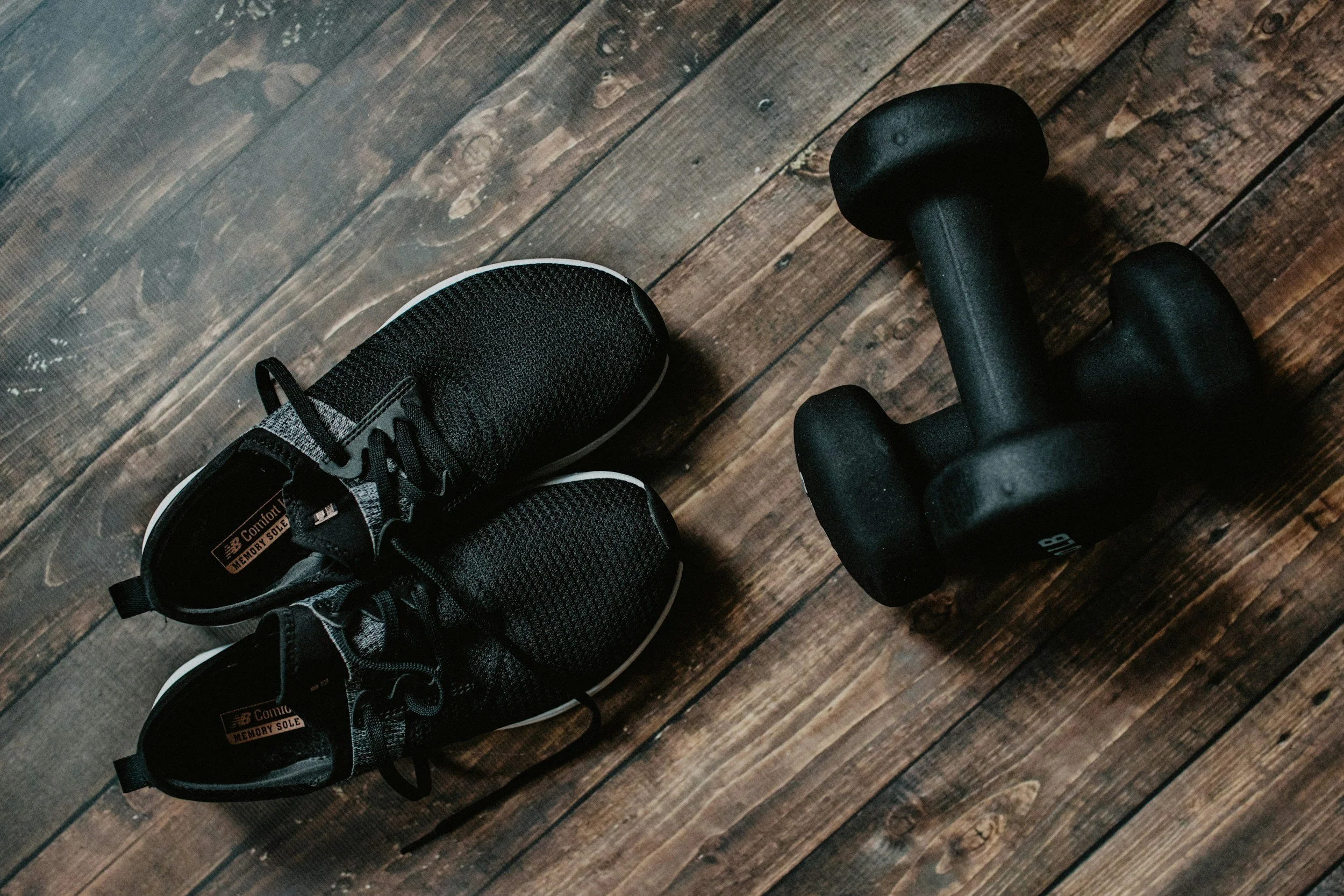 A pair of black athletic shoes, a black dumbbell, and a dark brown wooden floor.