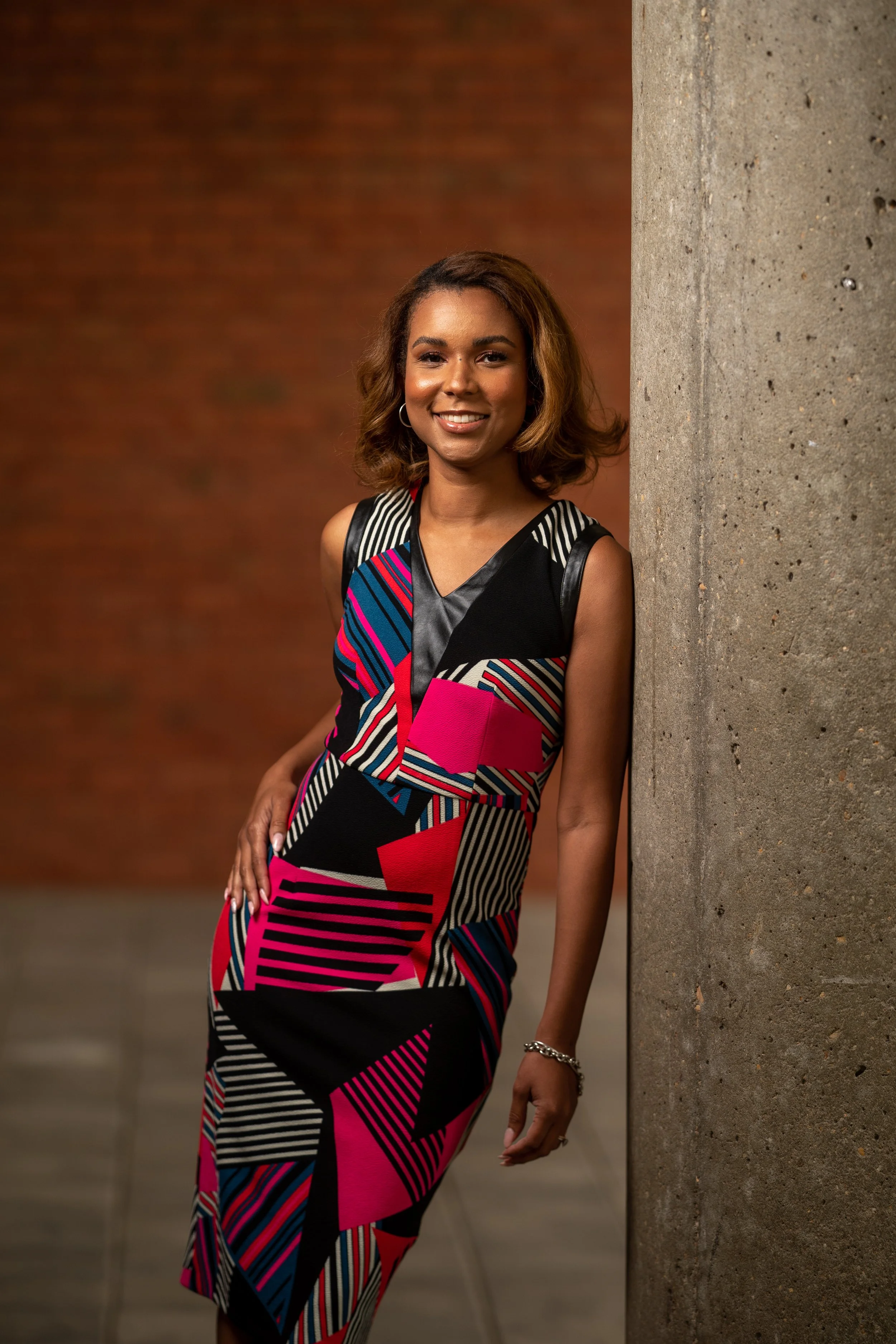 A woman with medium-length wavy hair, wearing a colorful sleeveless dress with a geometric pattern, standing next to a concrete wall, smiling at the camera.