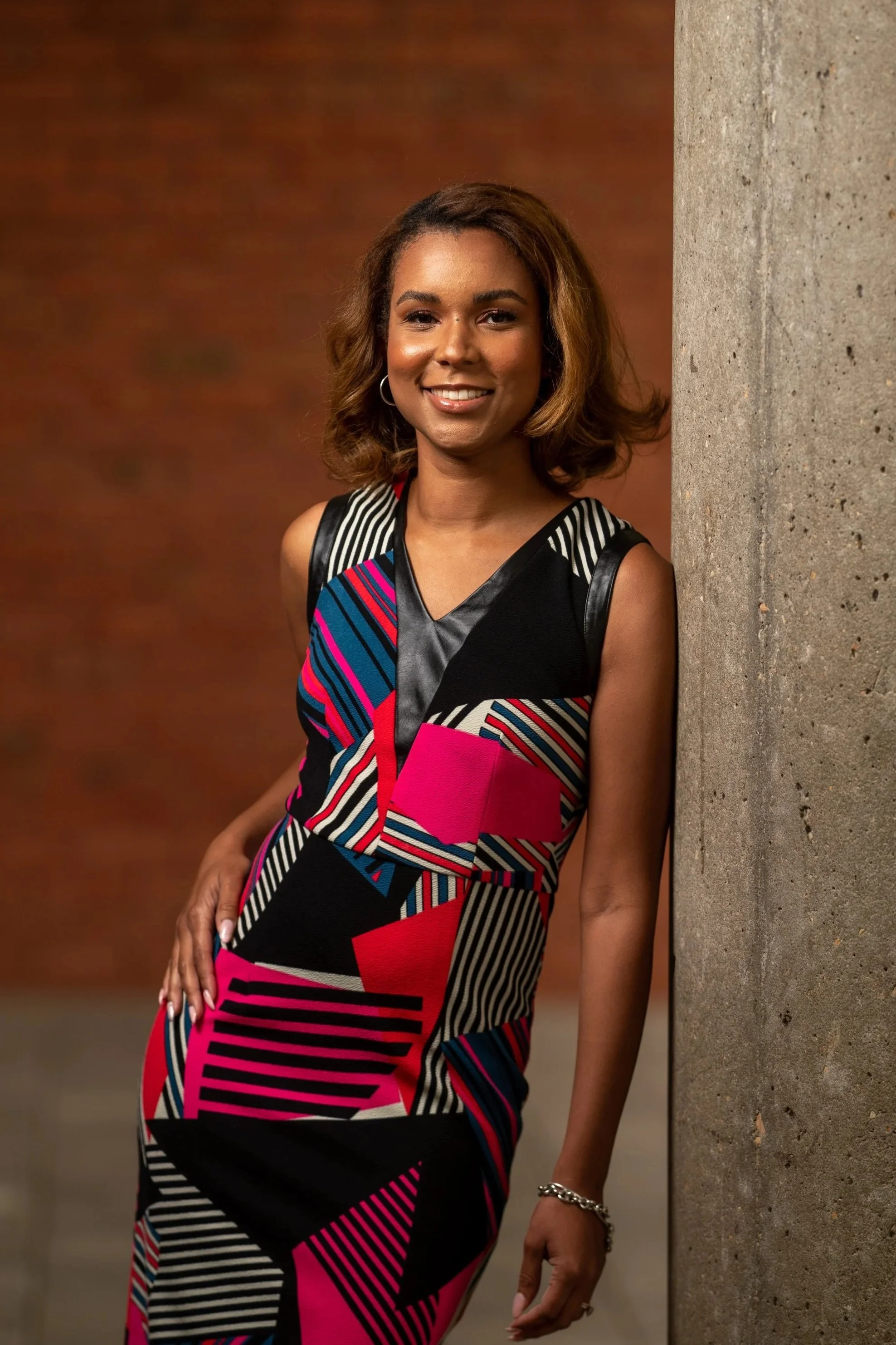 A woman with shoulder-length wavy hair, wearing a colorful geometric patterned dress, leaning against a concrete wall with a warm brown background.