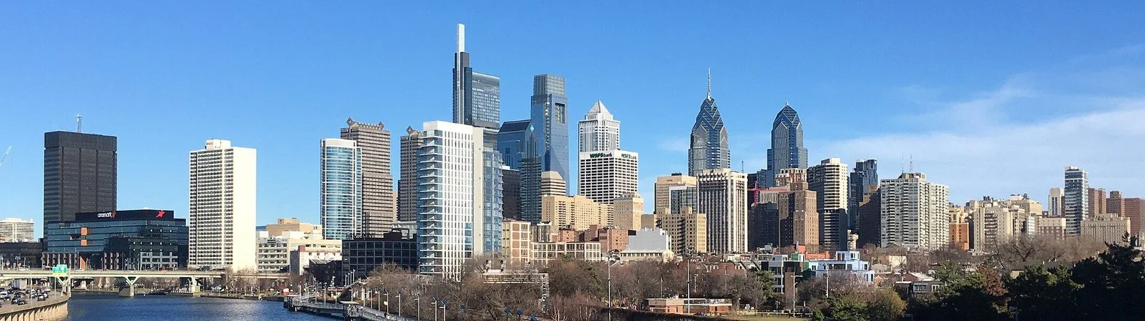 Panoramic view of downtown Philadelphia skyscrapers against a blue sky, with a river in the foreground.