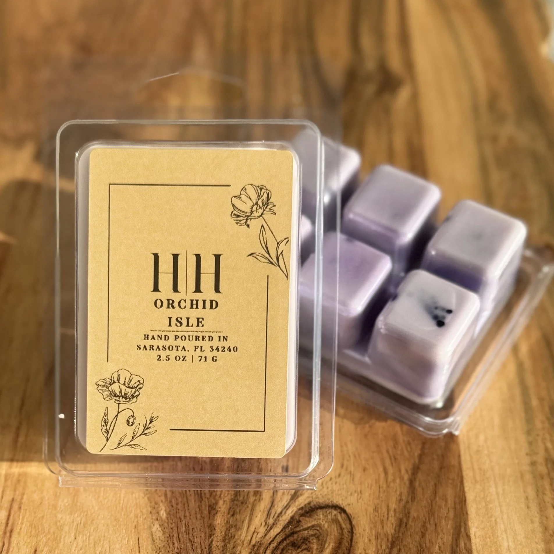 A clear plastic box with a beige label reading 'HH Orchid Isle, Hand Poured in Sarasota, FL, 2.5 oz, 71g' with floral illustrations, and several lavender-colored candles inside.