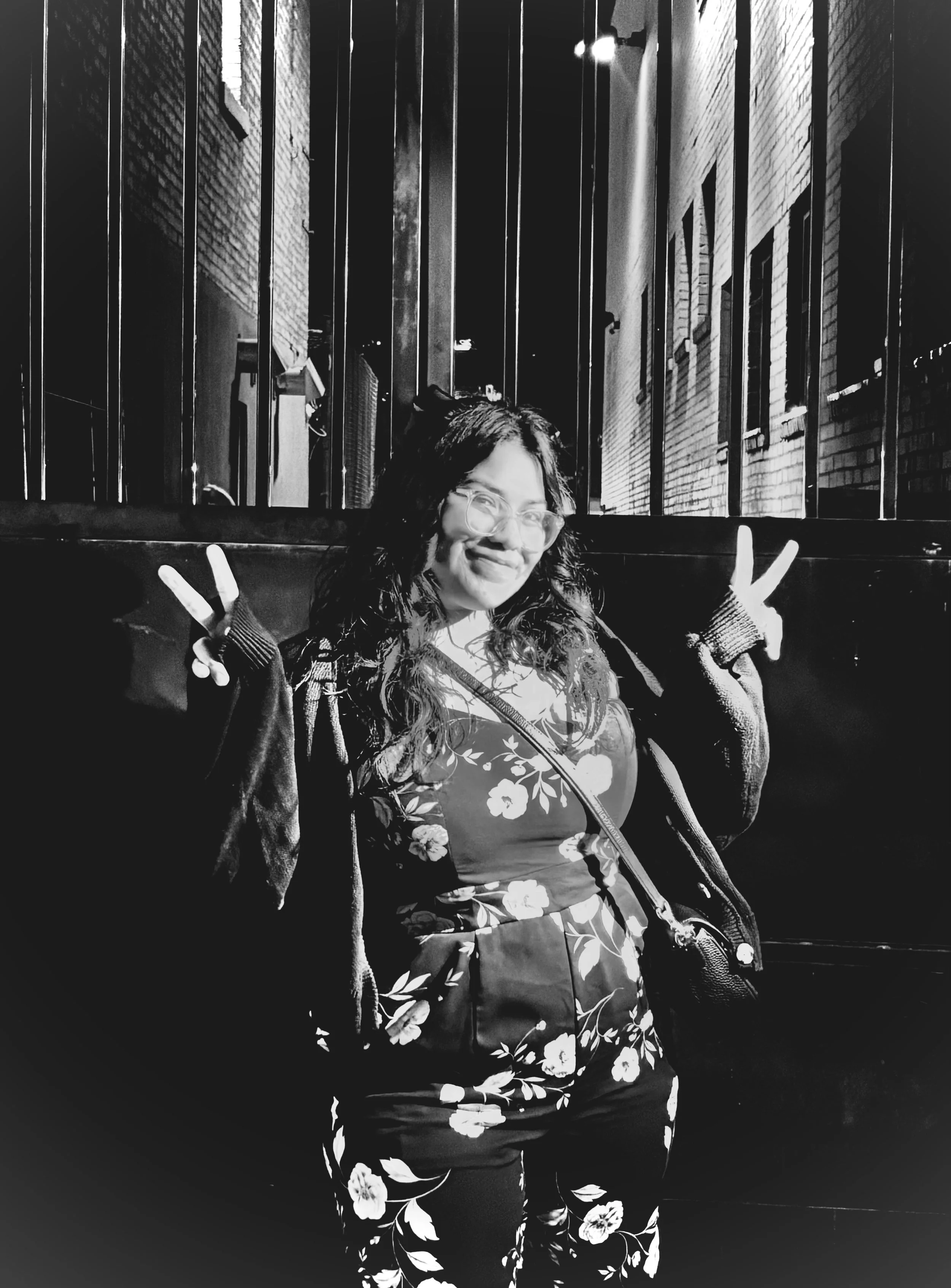 A woman with dark curly hair, glasses, and a floral dress posing with peace signs in a street setting at night with brick buildings.