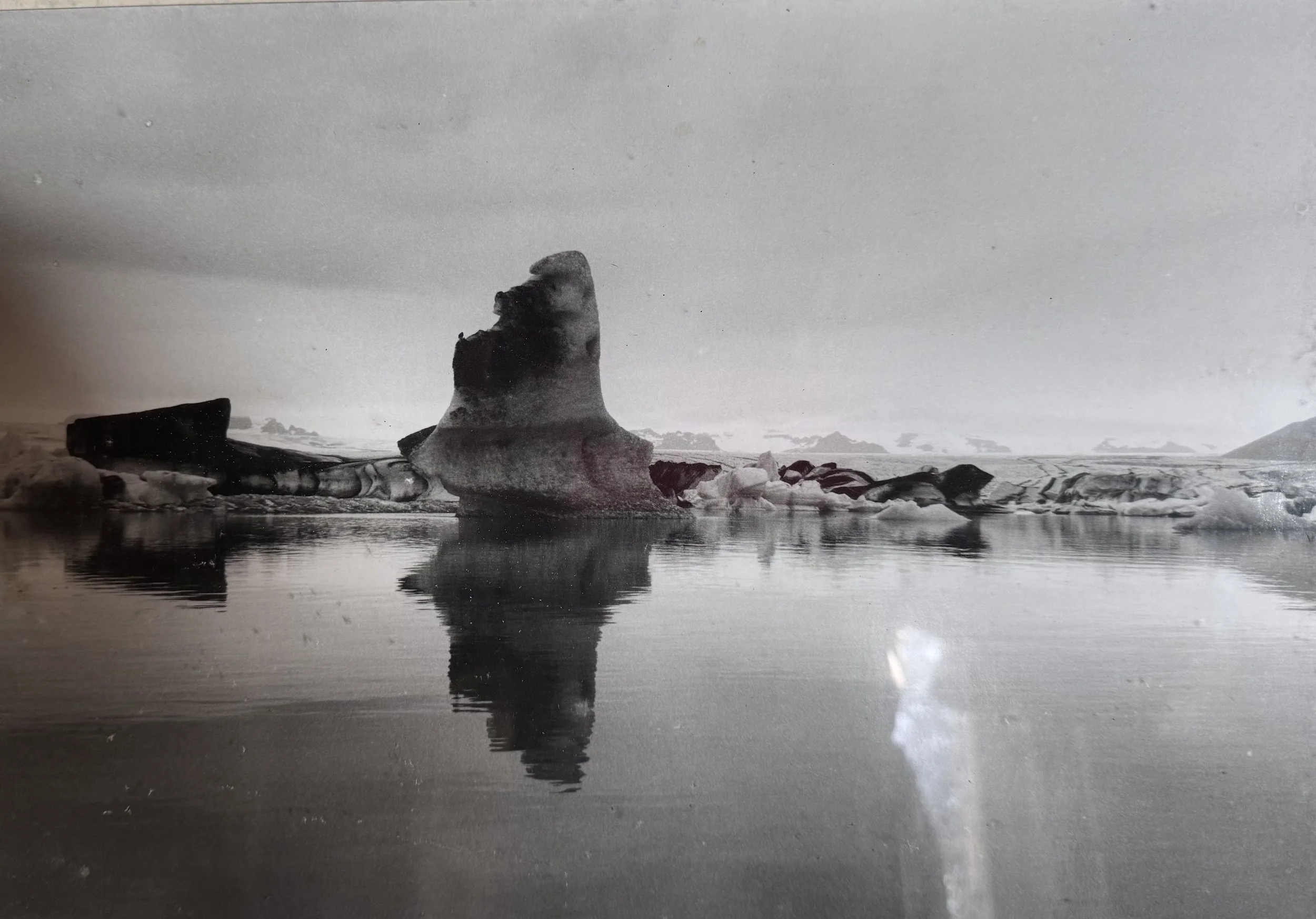Black and white photo of an iceberg floating in water, with a reflection visible on the water's surface.