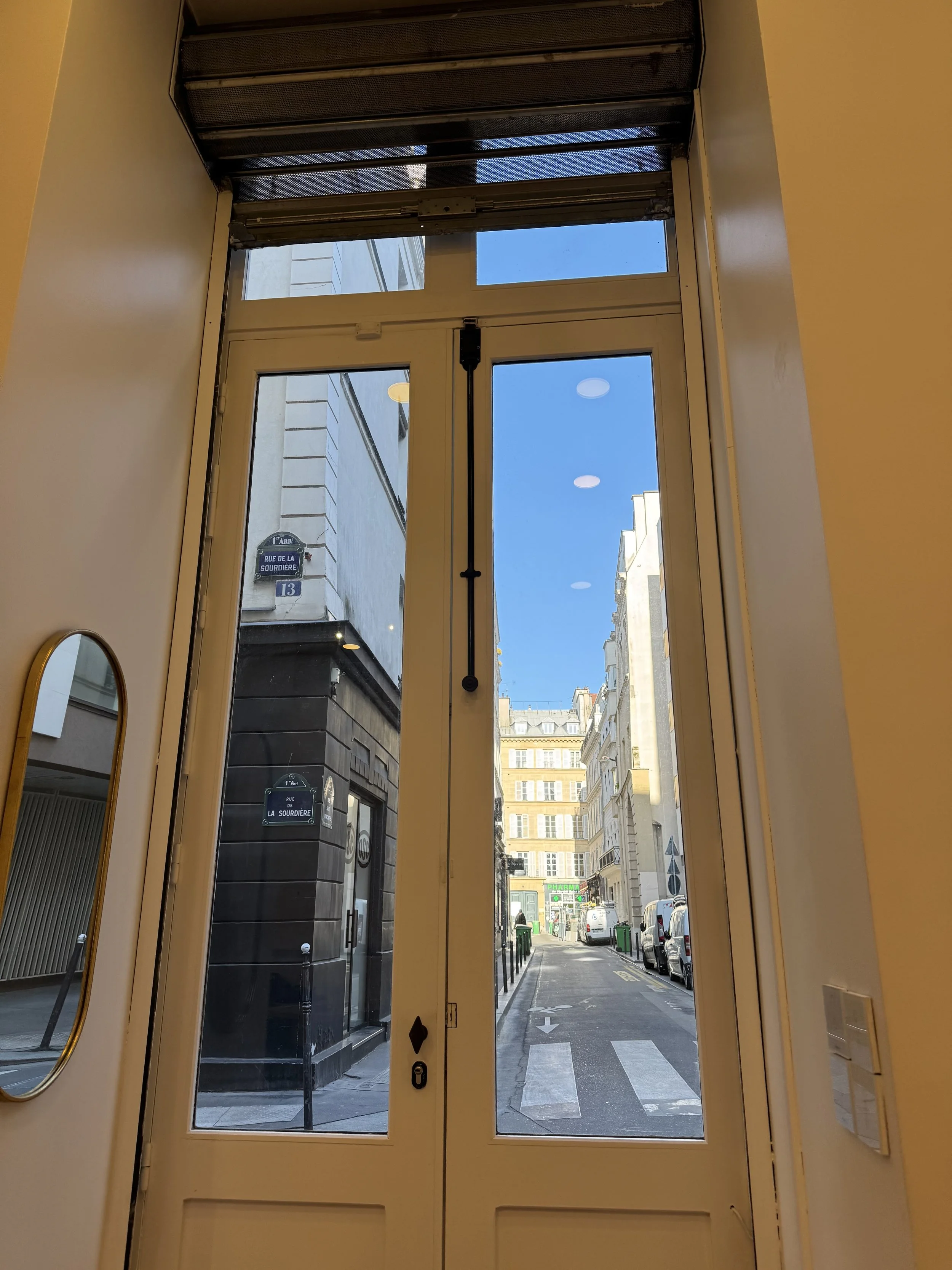 View of a city street through glass doors, with buildings, parked cars, and pedestrians, in a bright, sunny day in an urban area.