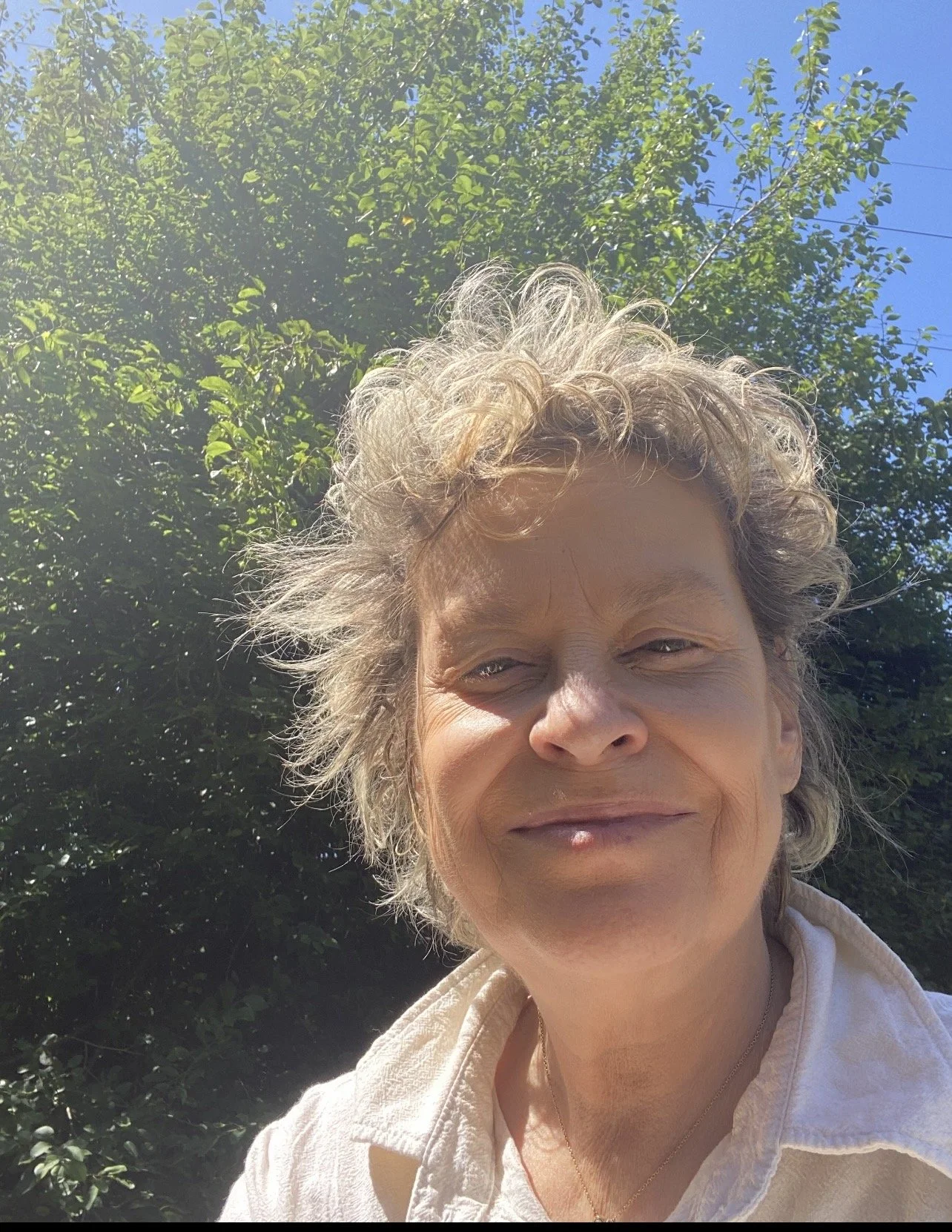 A woman with curly gray hair smiling outdoors, with trees and a clear blue sky in the background.