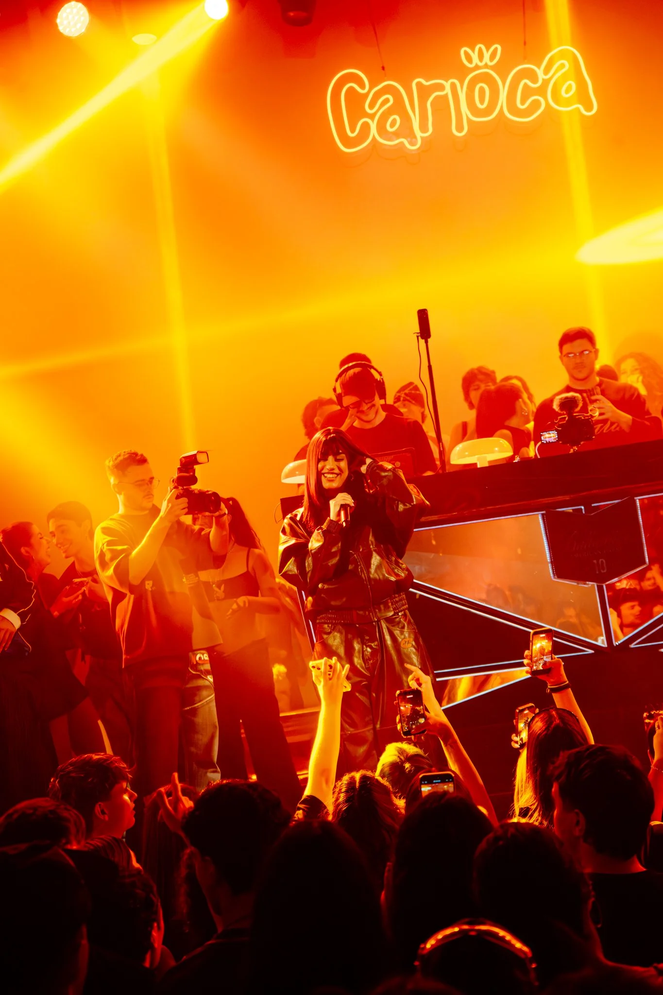 A singer in leather clothing performing on stage at a concert with a neon sign that says 'cárca' in the background, surrounded by an enthusiastic crowd taking photos and videos.
