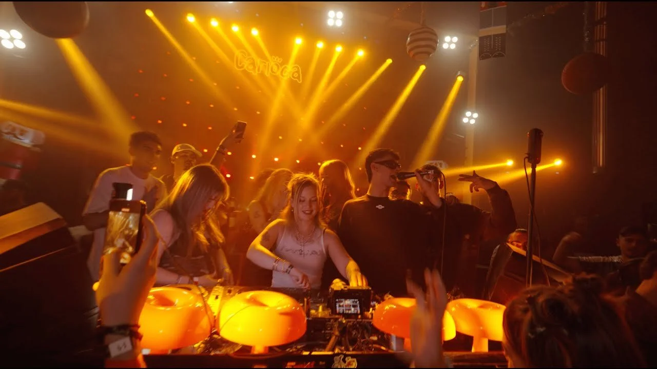 Two DJs perform at a music event with colorful stage lighting and sound equipment.