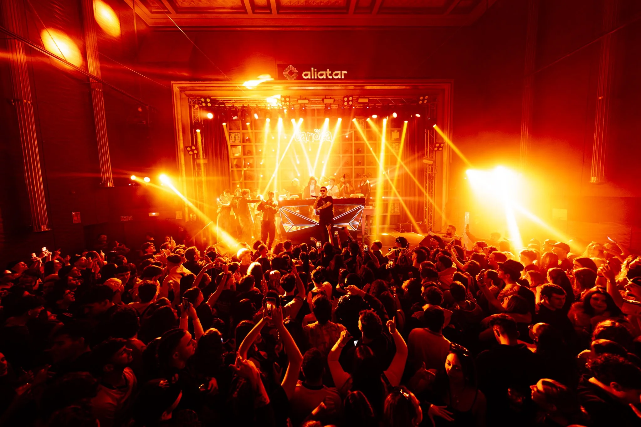 A DJ wearing a black jacket, white headphones around his neck, and a black New York Yankees cap, dancing and mixing music at a club with a red neon sign overhead.