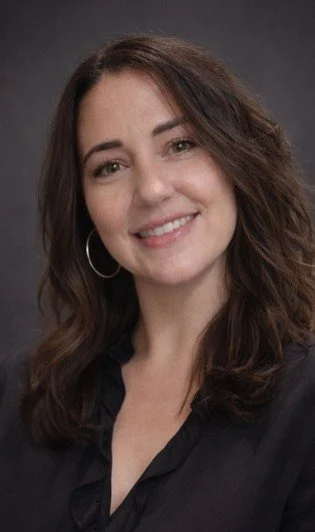 Portrait of a woman with long, wavy brown hair wearing hoop earrings and a black top, smiling at the camera.