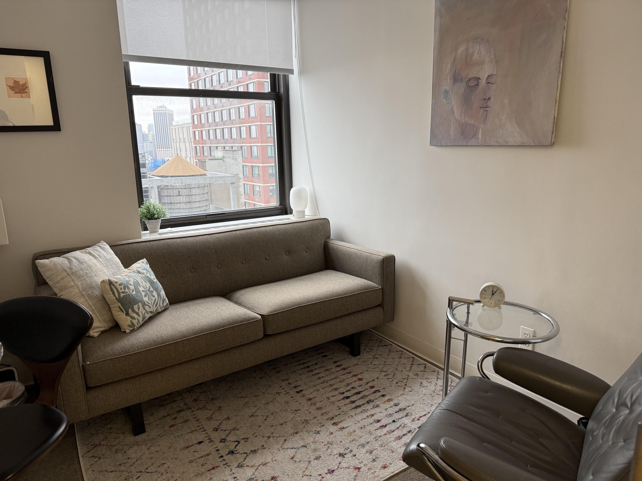 Therapy office room space with beige sofa, window showing cityscape, abstract portrait on wall, glass side table with clock, leather chair, and hanging picture. Cozy space to meet with a therapist in person in Downtown Brooklyn.