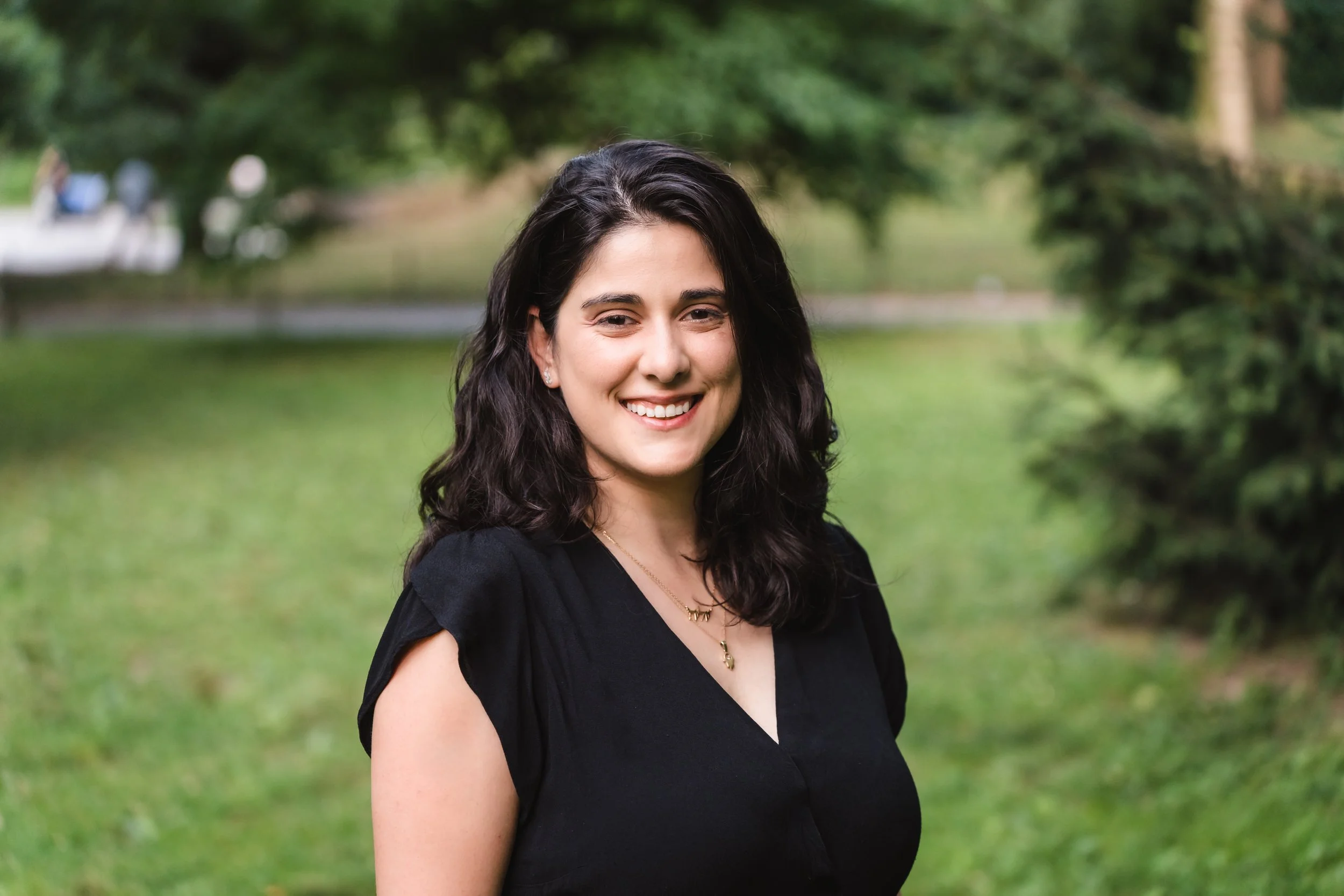 A woman with shoulder-length dark hair smiling outdoors in a park, wearing a black top and gold jewelry.