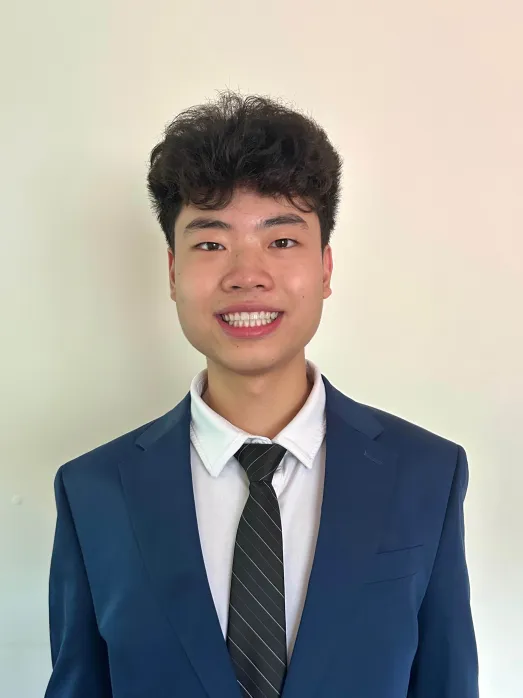 Young man smiling, wearing a navy suit, white shirt, and black striped tie in front of a plain off-white wall.