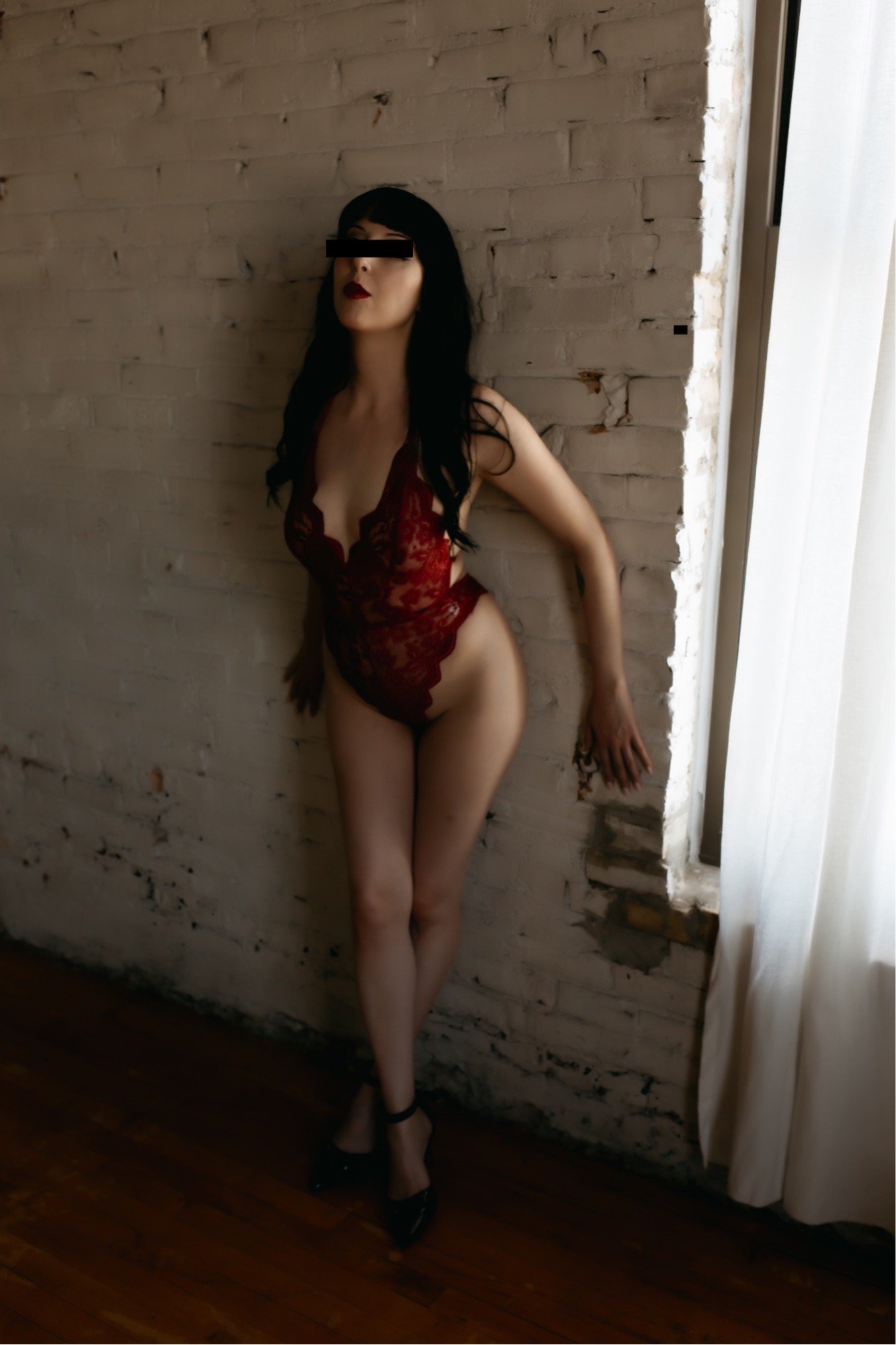 A woman with black hair and red lipstick wearing a red lace bodysuit and black high heels, standing against a white brick wall near a window with curtains.