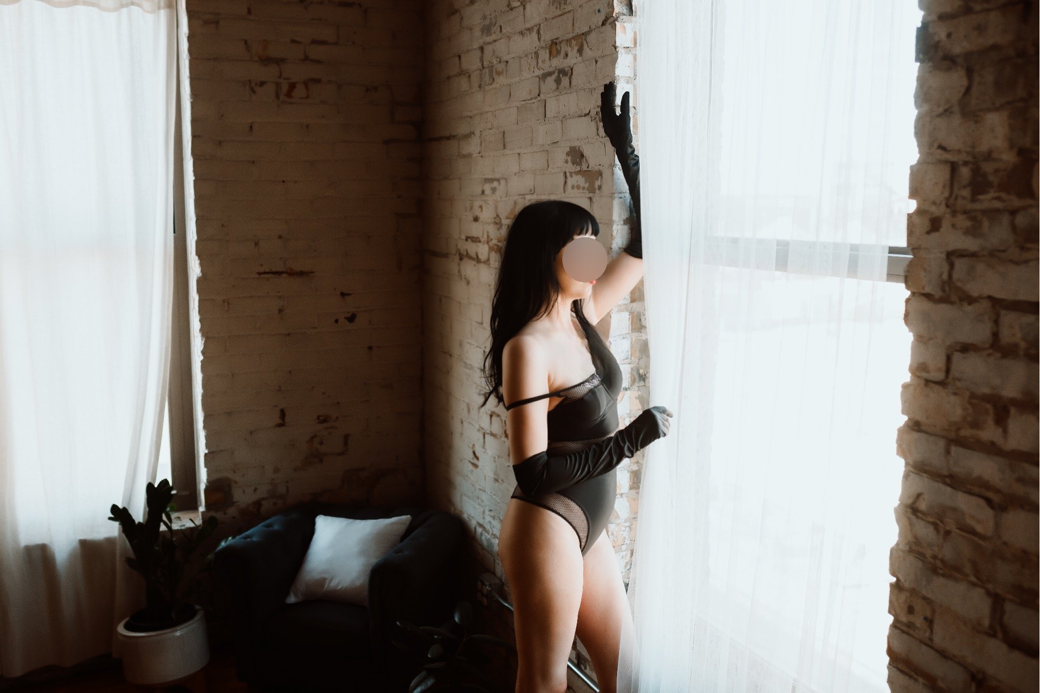 A woman in black lingerie and long black gloves standing near a window with sheer curtains, with a brick wall behind her.