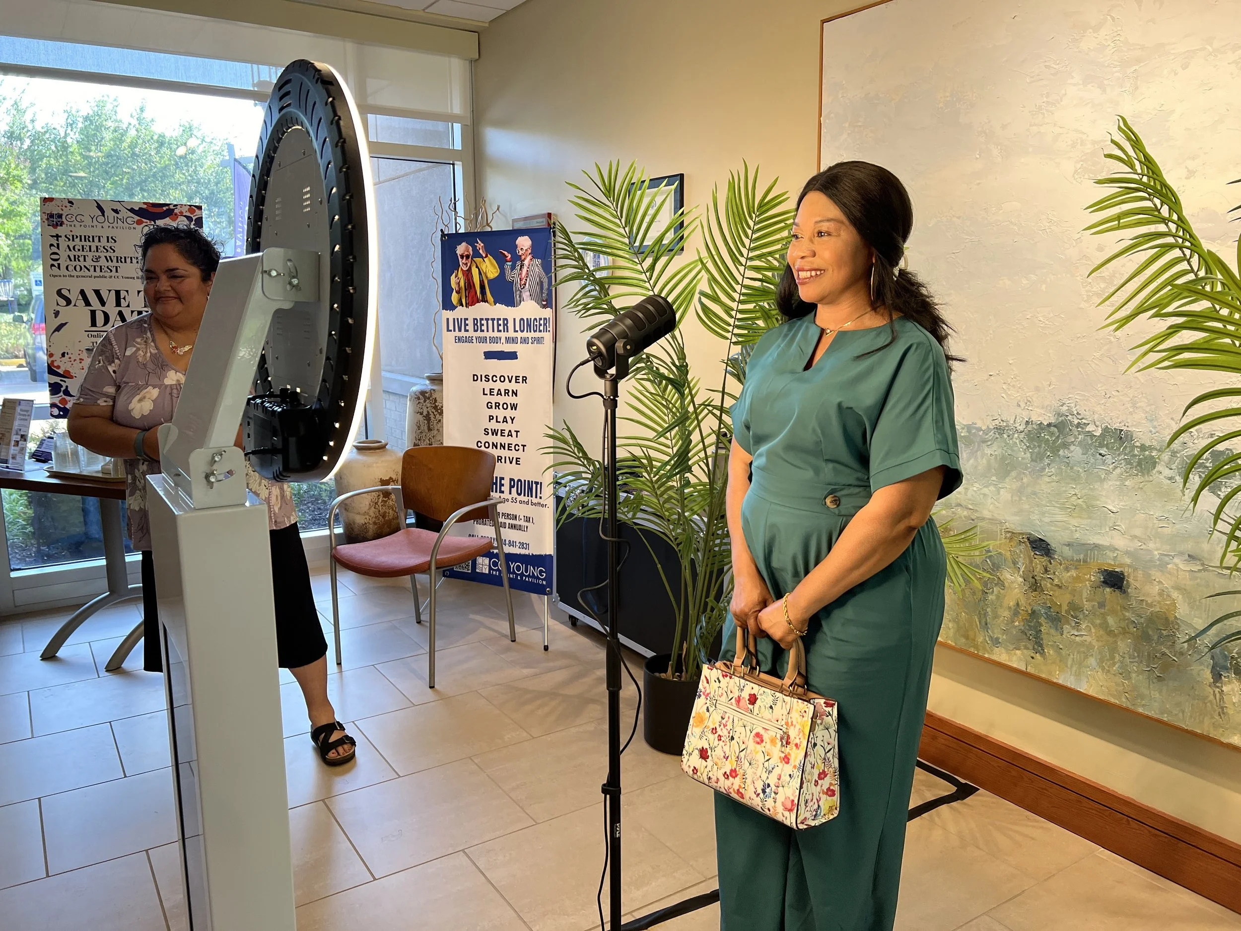 A woman in a green dress is smiling and speaking into a microphone using the DFW Booths Video Guest Book, with decorative plants as a backdrop behind her.