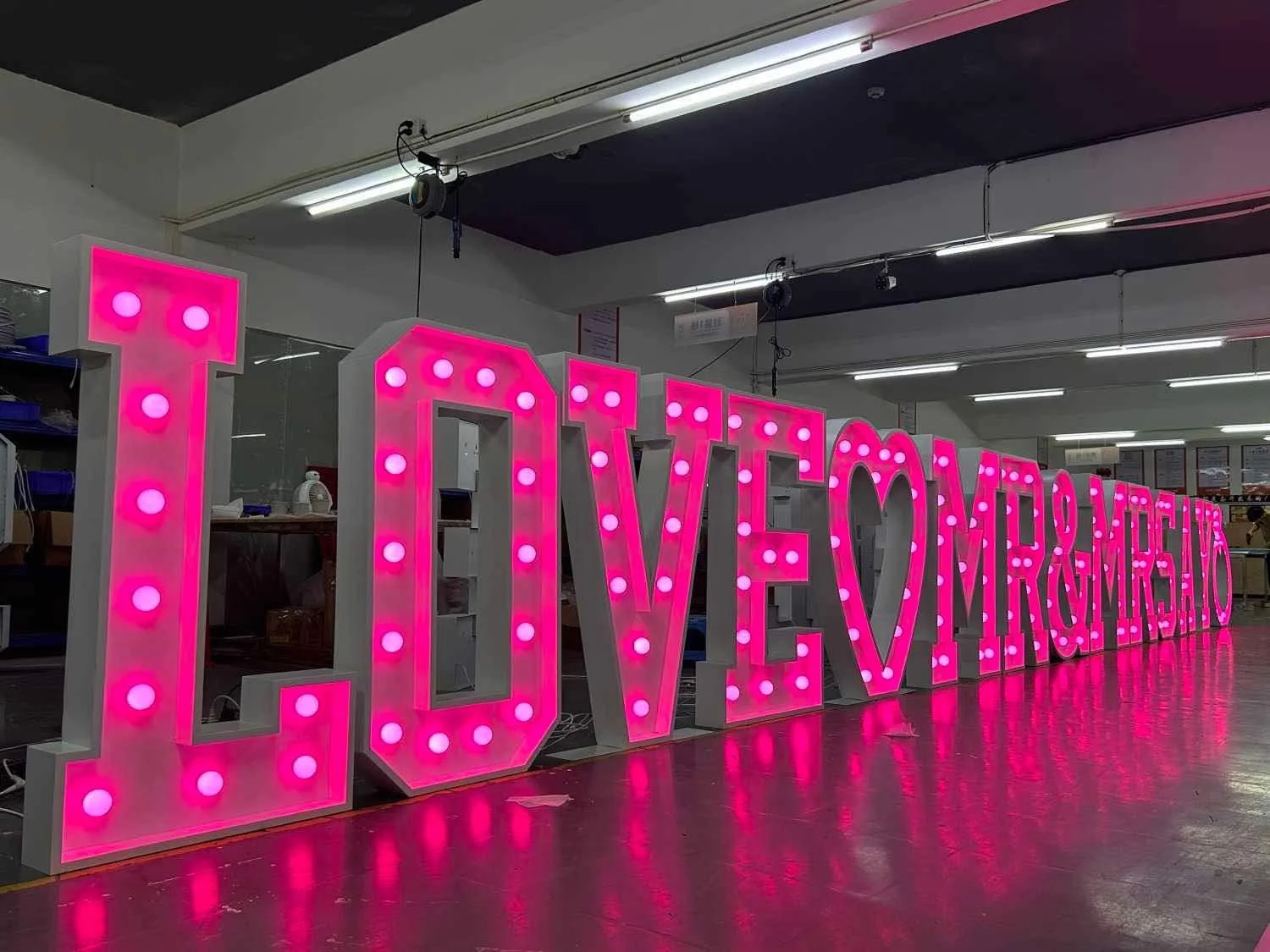 Large illuminated marquee letters spelling the words "LOVE" and "MR. AND MRS." in big pink letters with white bulbs, inside a store or warehouse with overhead fluorescent lighting.
