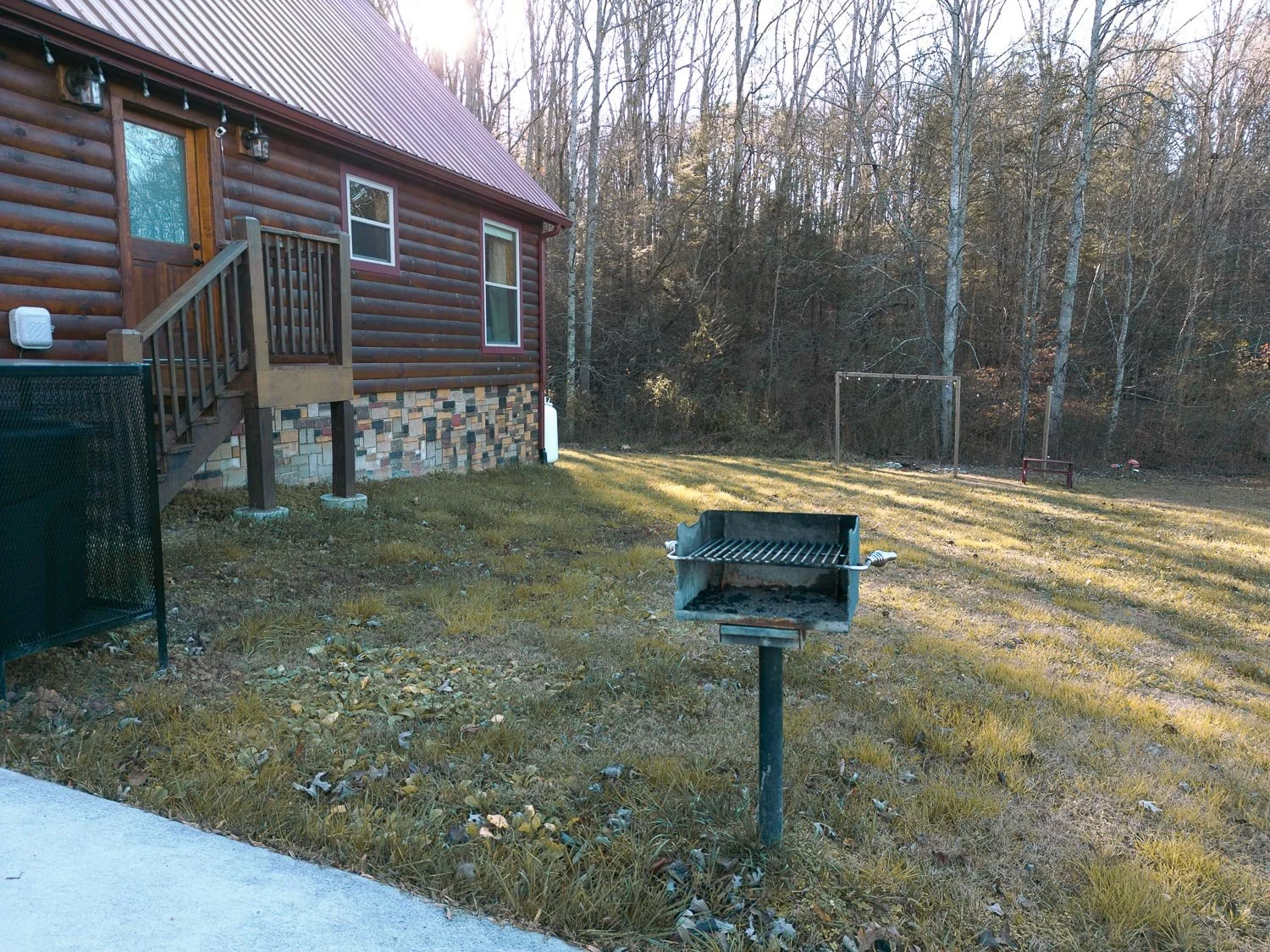 Backyard with a small wooden deck attached to a log cabin, a charcoal grill, a soccer goal, and leafless trees in the background.