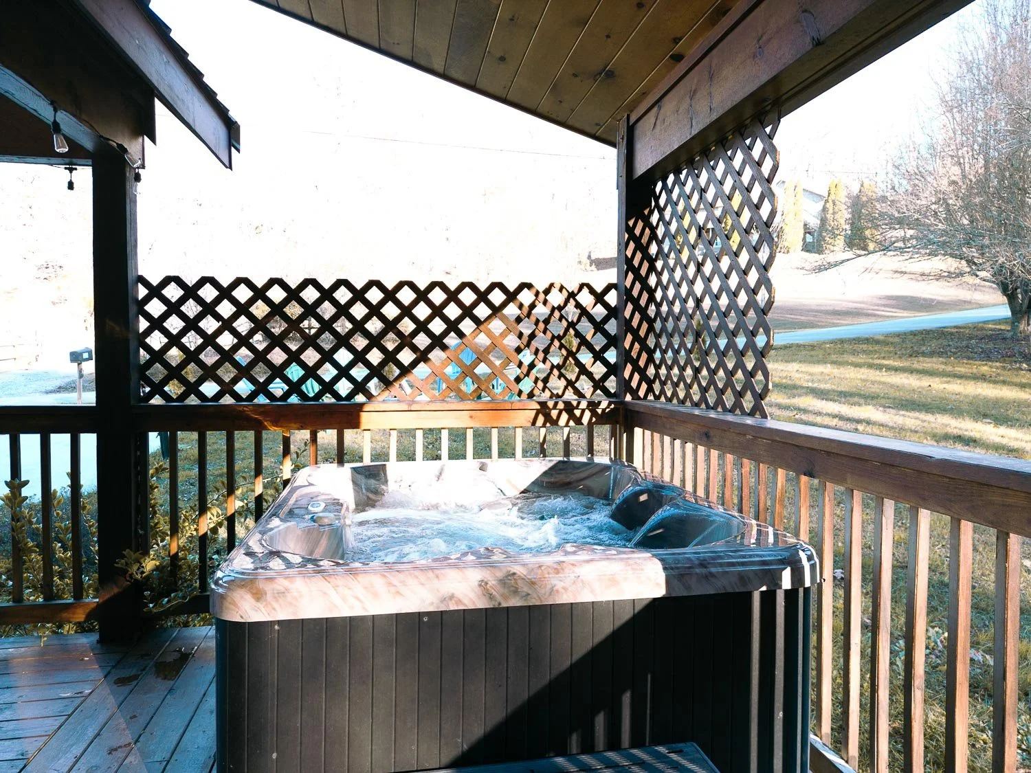 Hot tub on a wooden porch with lattice side panels, overlooking a yard and a leafless tree during daytime.