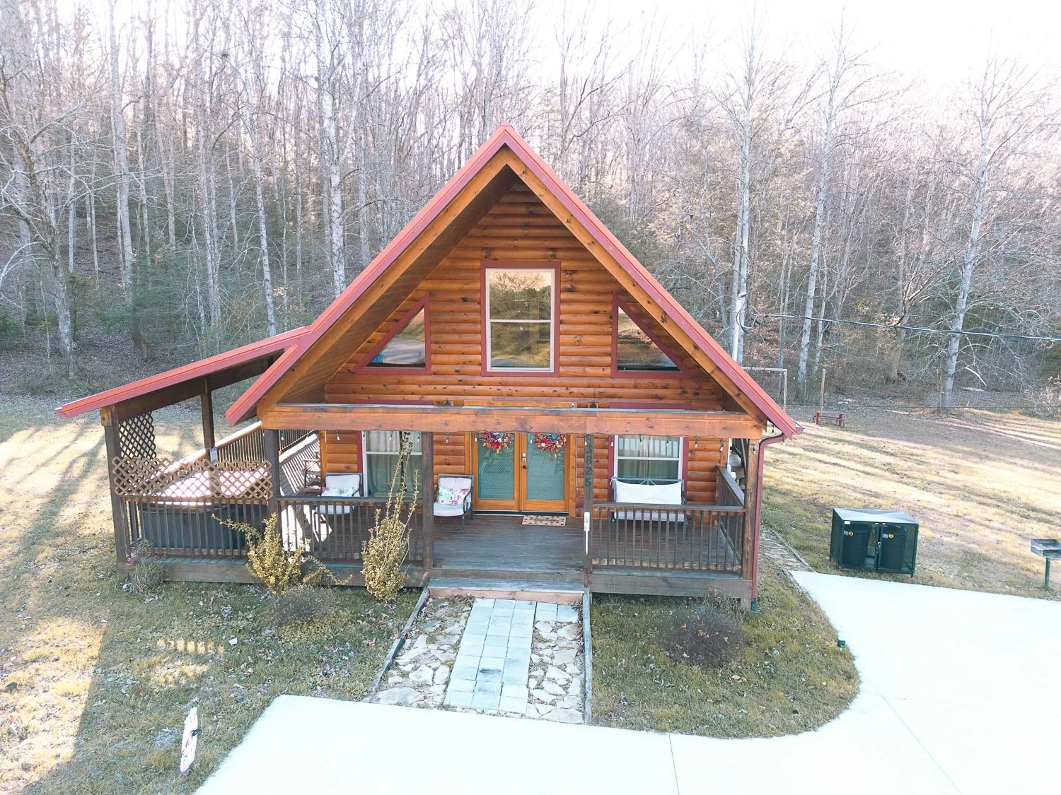 A two-story wooden house with a large front porch, decorated with flower baskets and outdoor furniture, surrounded by a grassy yard and located near a wooded area with tall leafless trees.