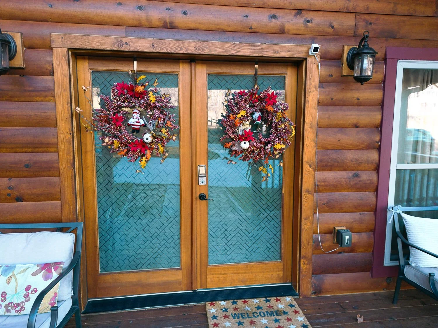 Decorated wooden front door with two holiday wreaths, outdoor seating on porch, welcome mat, and exterior wooden wall with a lantern-style light fixture.