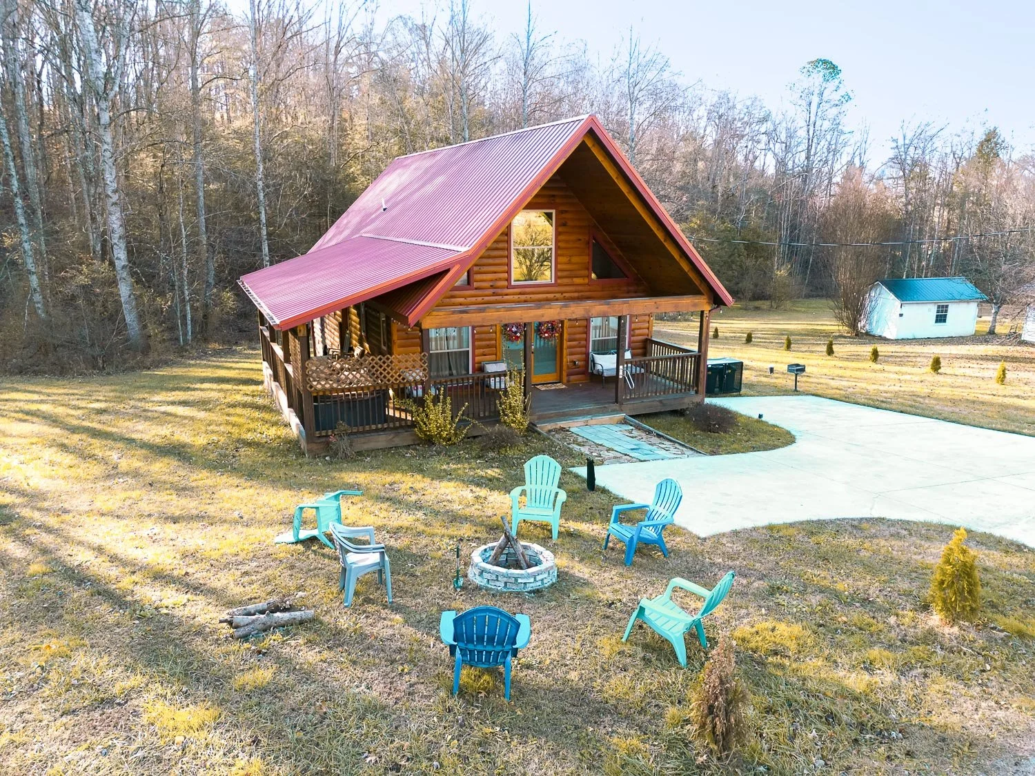 A wooden log cabin with a brown roof and a front porch surrounded by a grassy yard with outdoor seating and a fire pit, set near a wooded area with leafless trees.