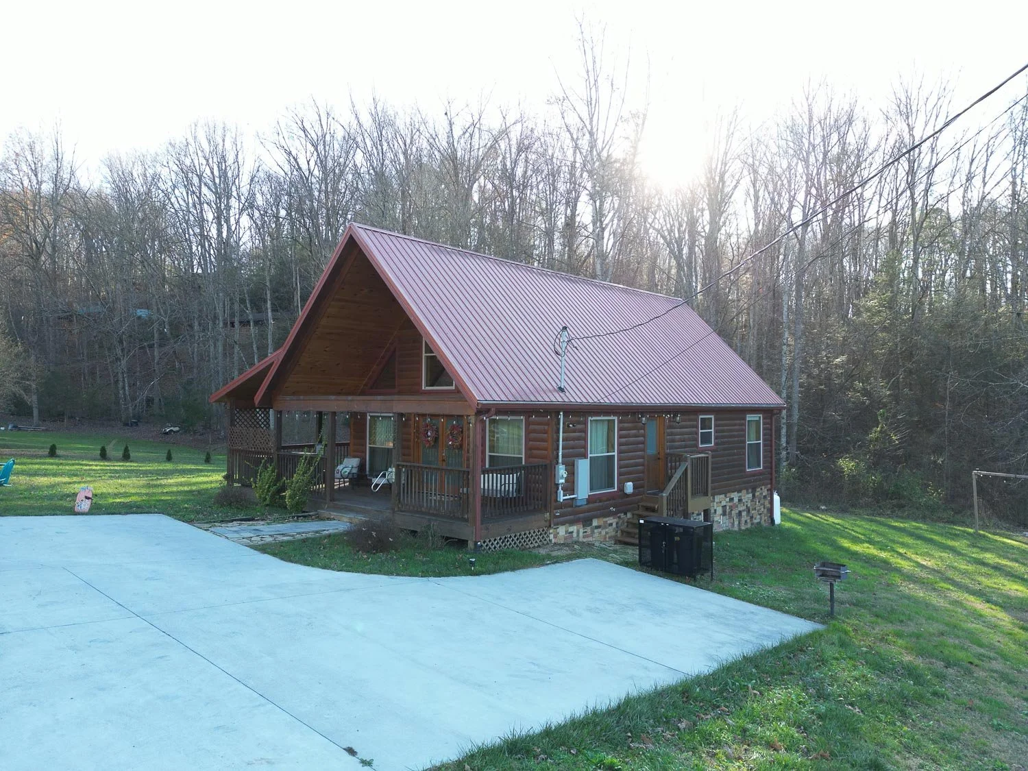 A wooden house with a red metal roof and a front porch, set in a grassy yard with a concrete driveway, trees in the background, and the sun shining overhead.