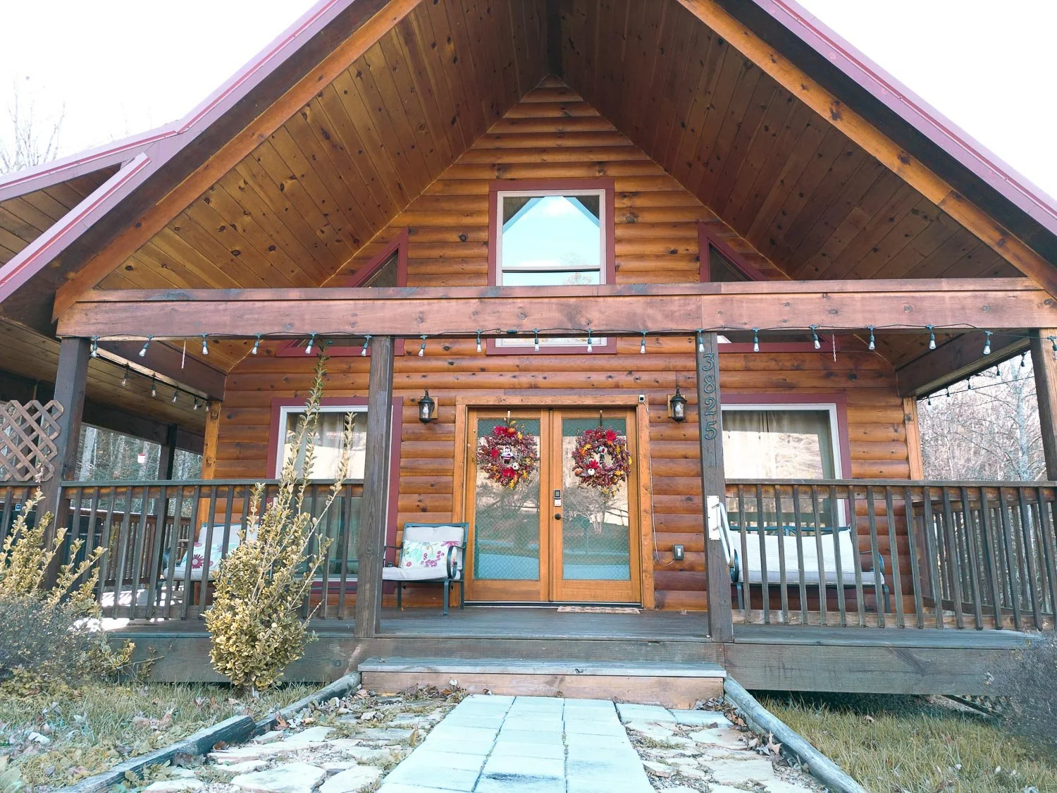 Wooden house with front porch decorated with two wreaths on the double doors, two benches, string lights, and plants in the yard.