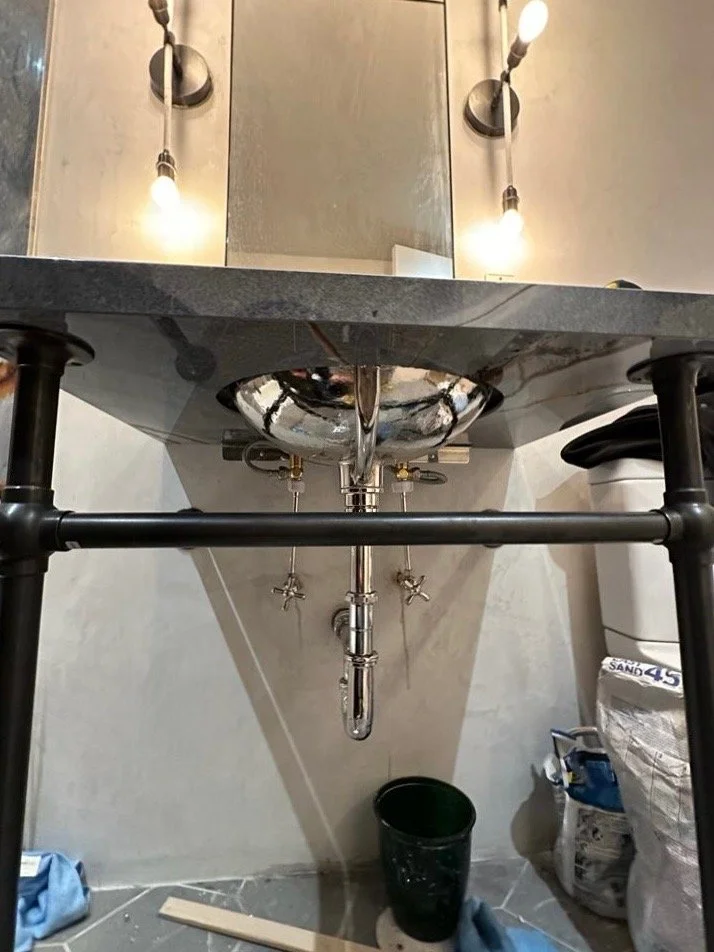 A stainless steel sink with a pipe underneath, mounted on a black metal stand. To the right, there is a bag of sand and a toilet. A black bucket and blue cleaning cloth are on the floor. Two wall-mounted light fixtures with exposed bulbs are above th