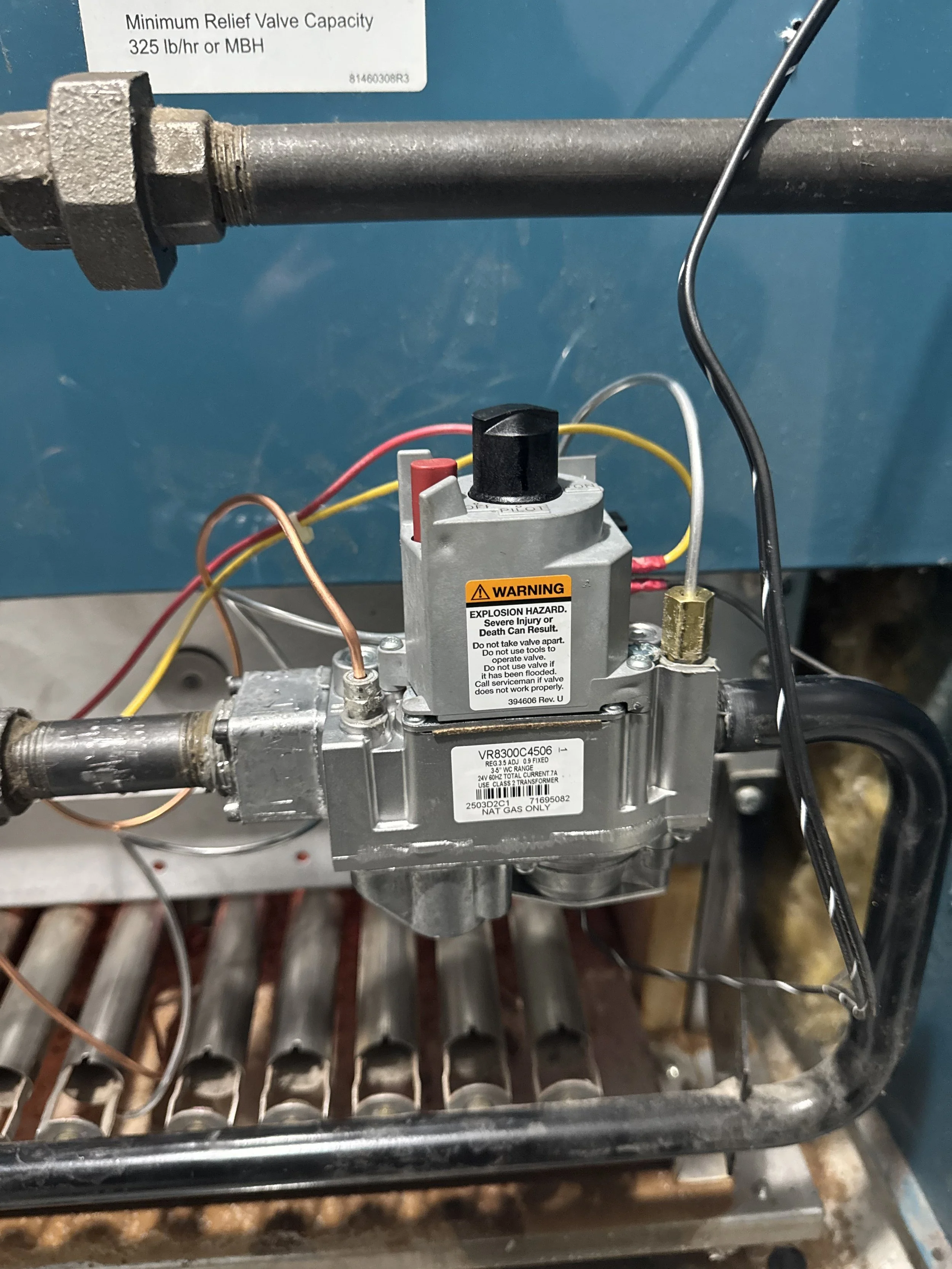 A close-up of a mechanical gas valve assembly with wires connected, mounted on a metal frame, with pipe fittings attached on either side, and a label indicating a minimum relief valve capacity of 325 lbs/hr or MBH.