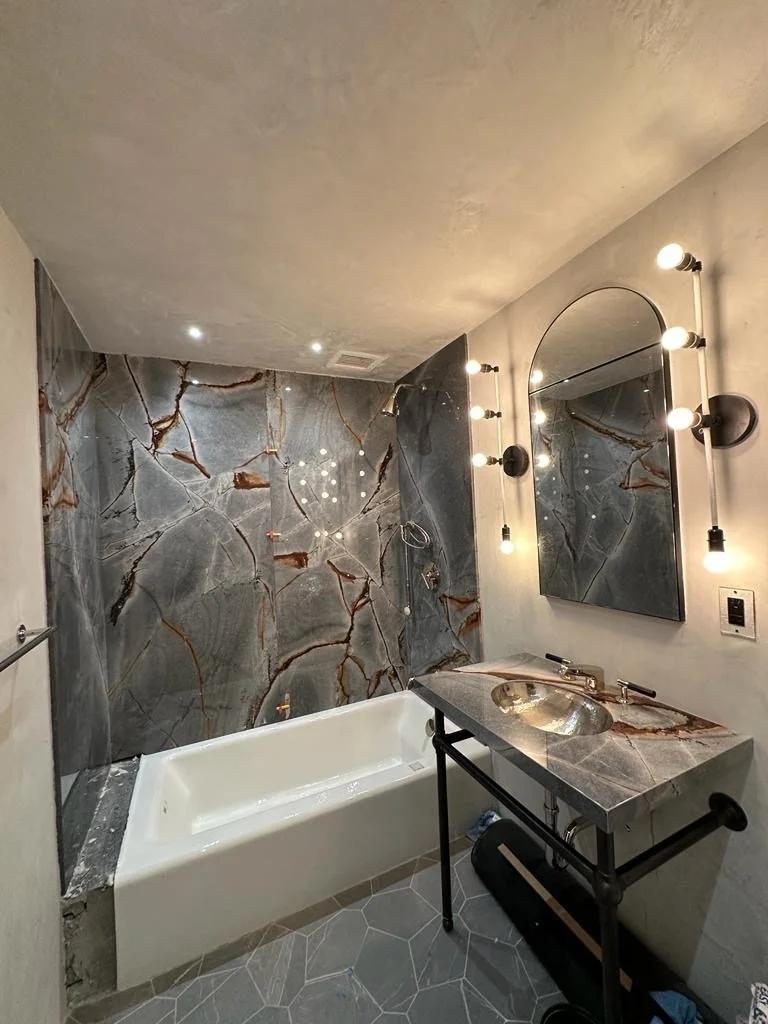 Modern bathroom with a bathtub, a marble vanity with a sink, a large arched mirror, and wall-mounted light fixtures. The wall behind the bathtub is decorated with large marble tiles with brown veining.
