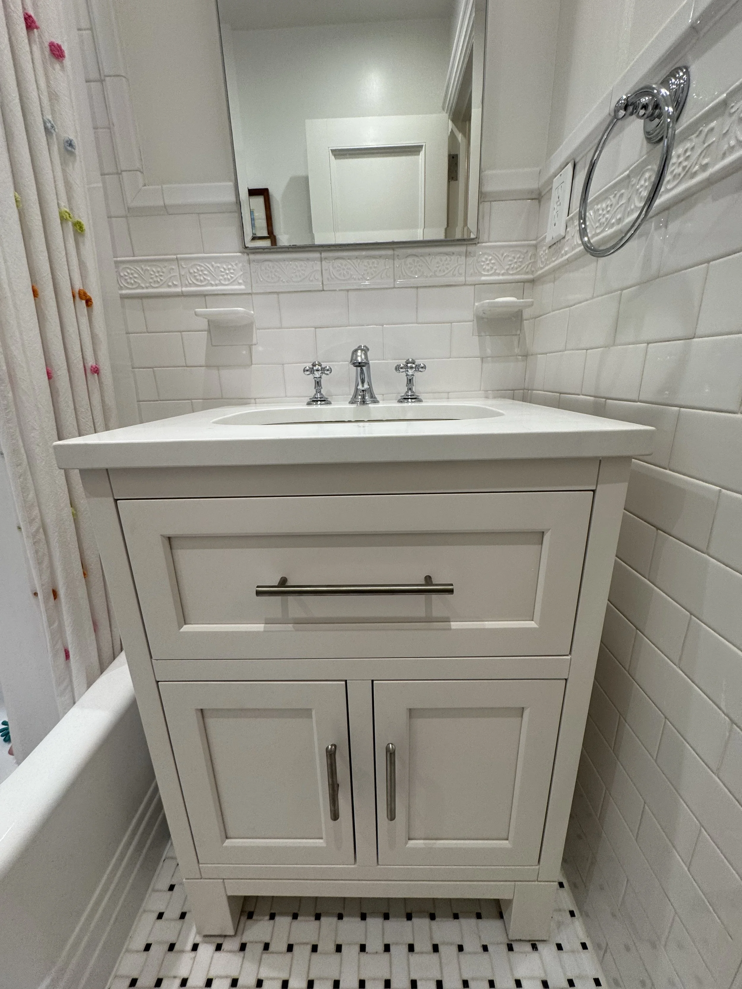 A white bathroom vanity with a rectangular sink, two cabinet doors, a drawer, and a mirror above it in a bathroom with white tiled walls.