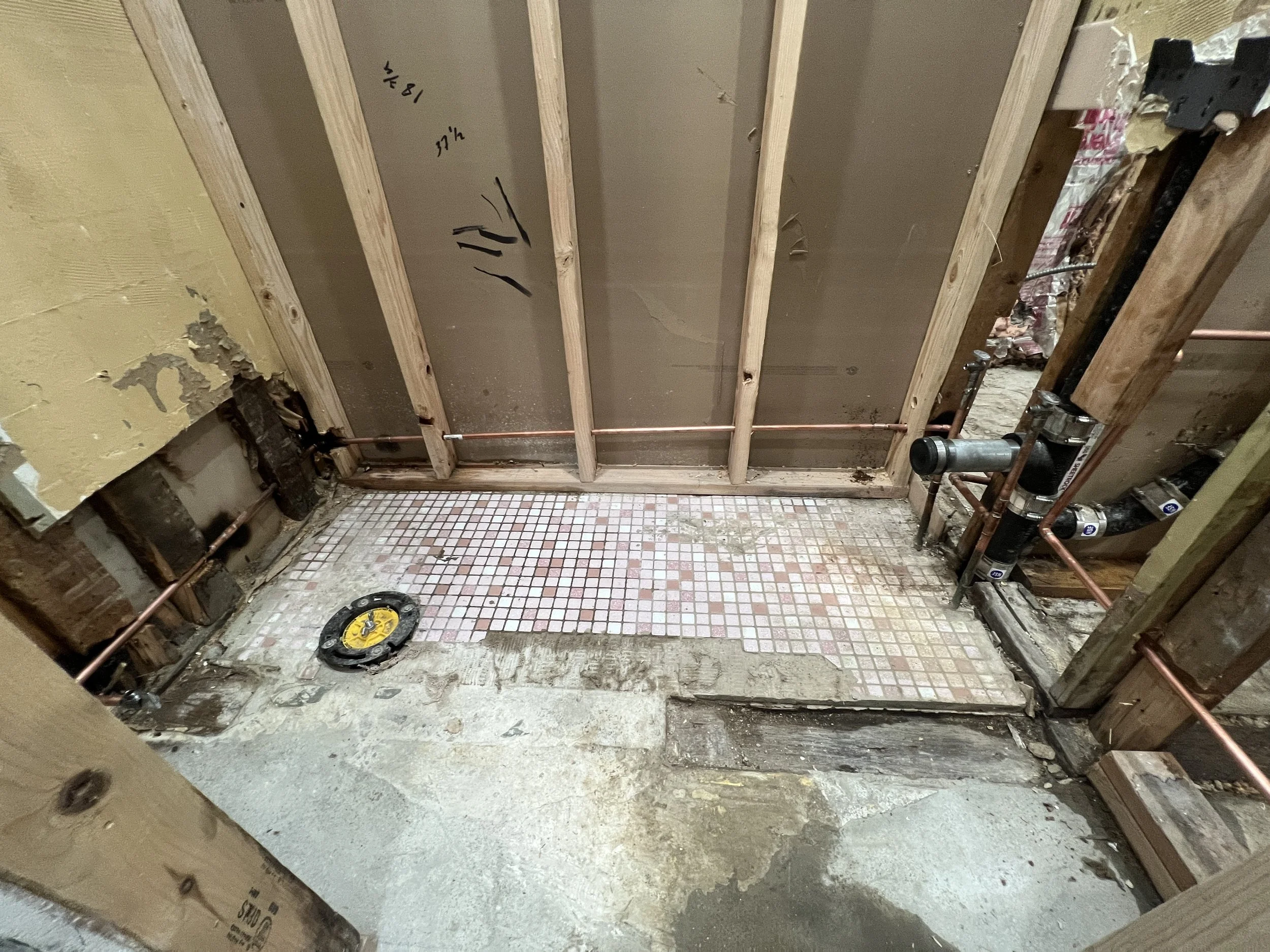 Construction site showing plumbing pipes and unfinished walls with wooden framing and small tiled floor area.