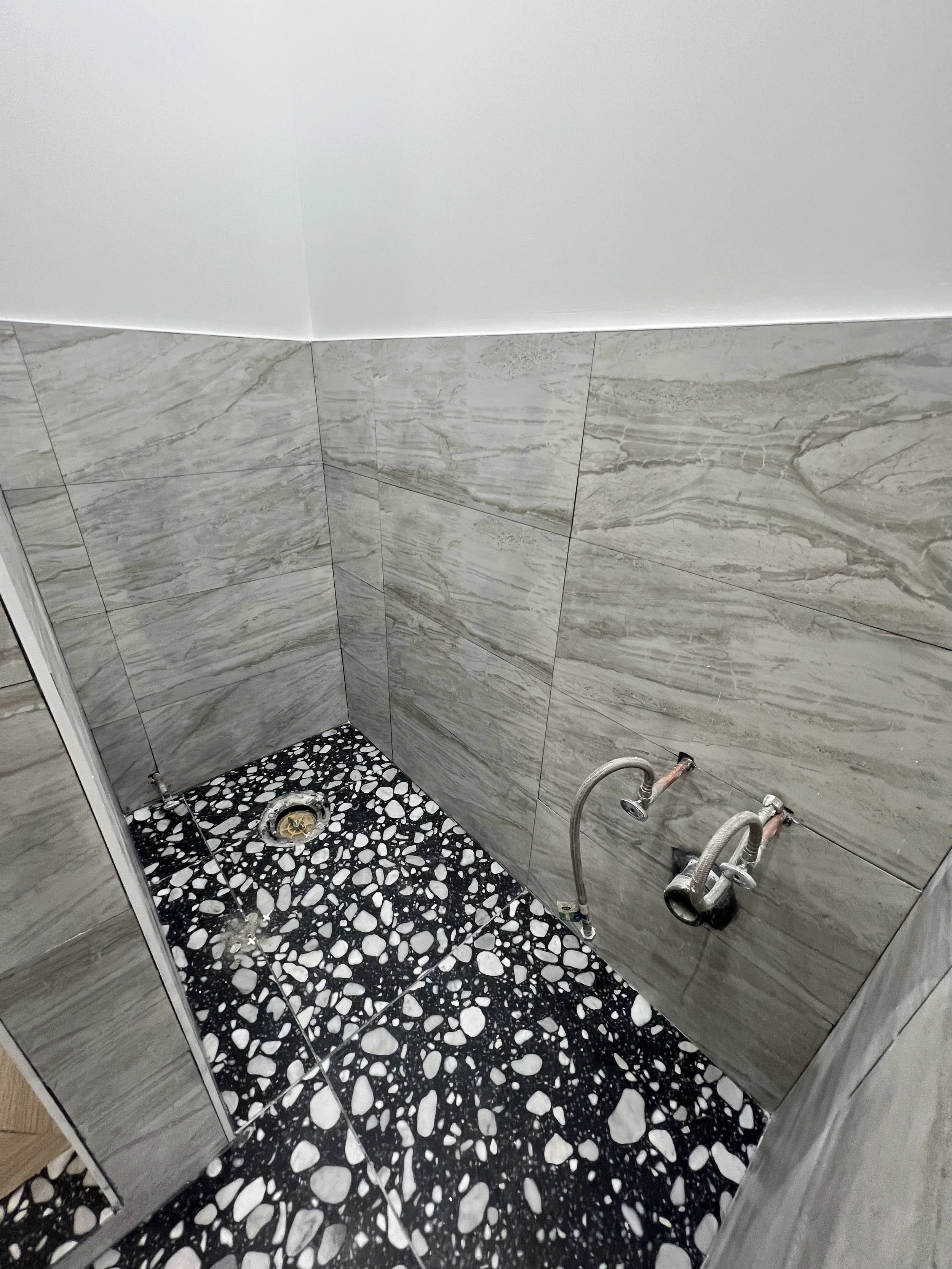 Empty shower area with marble tiles on the walls and black and white pebble-patterned tiles on the floor, with exposed pipes and drain.