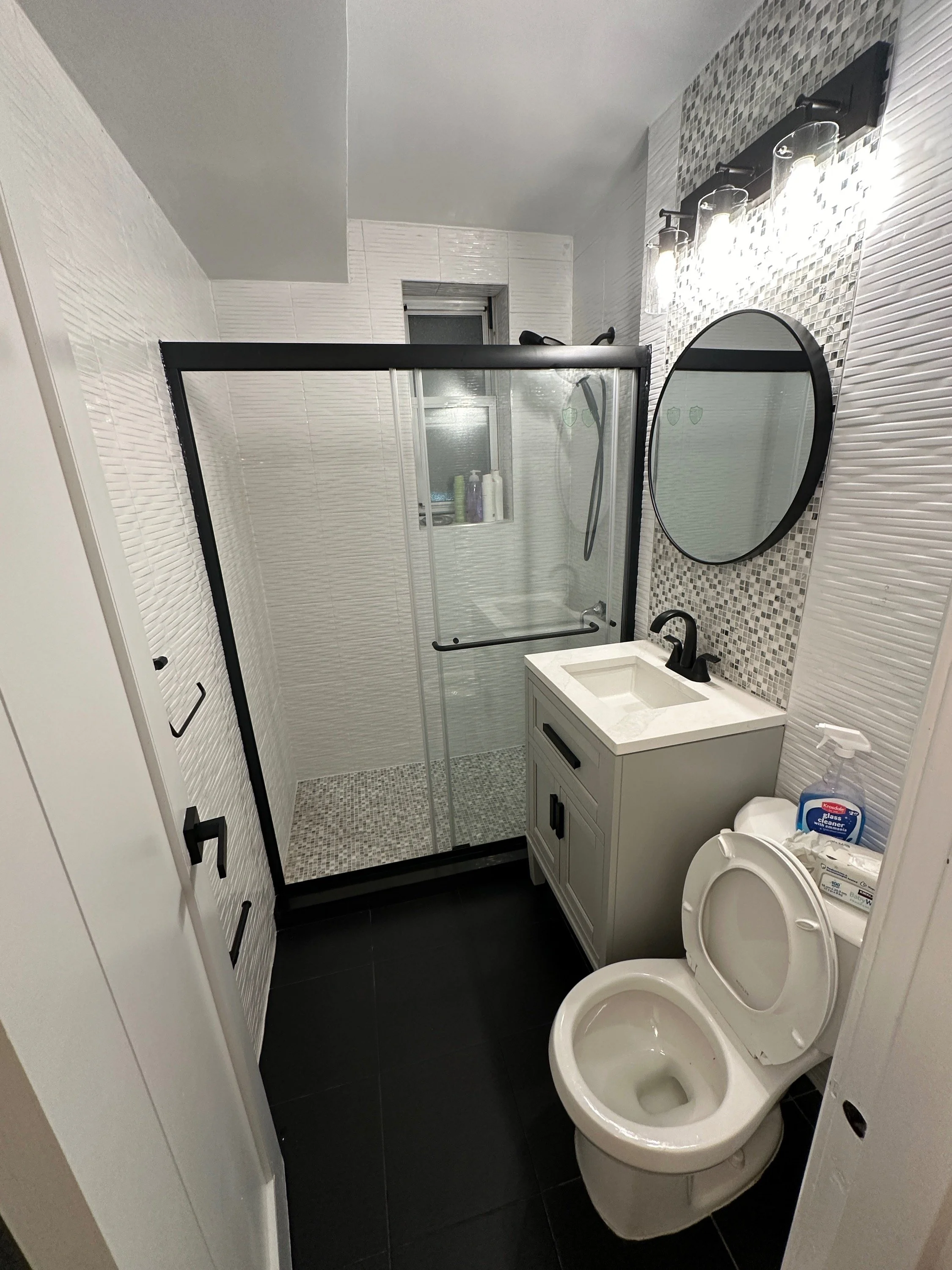 A small modern bathroom with a standing glass shower, white vanity with a round mirror, toilet, and black fixtures, decorated with white textured subway tiles and small mosaic tiles.