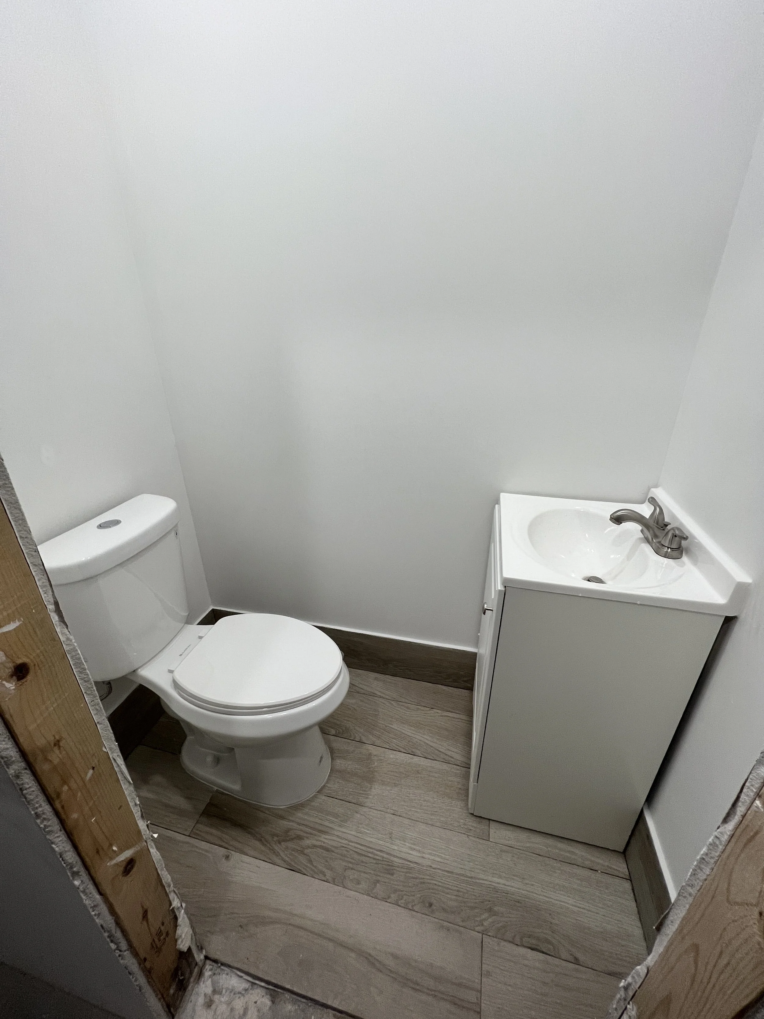Small bathroom with a toilet and a white sink vanity in a corner, wood-look flooring, plain white walls, and unfinished door frame.