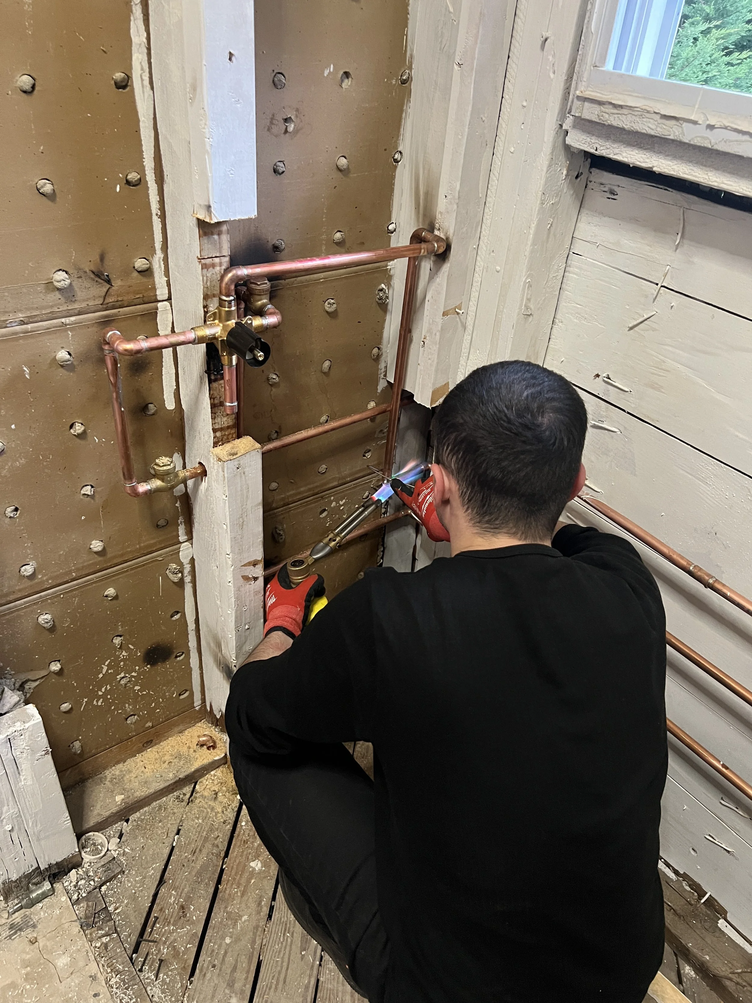 A person welding copper pipes inside a construction or renovation site, with exposed wooden wall and a window.