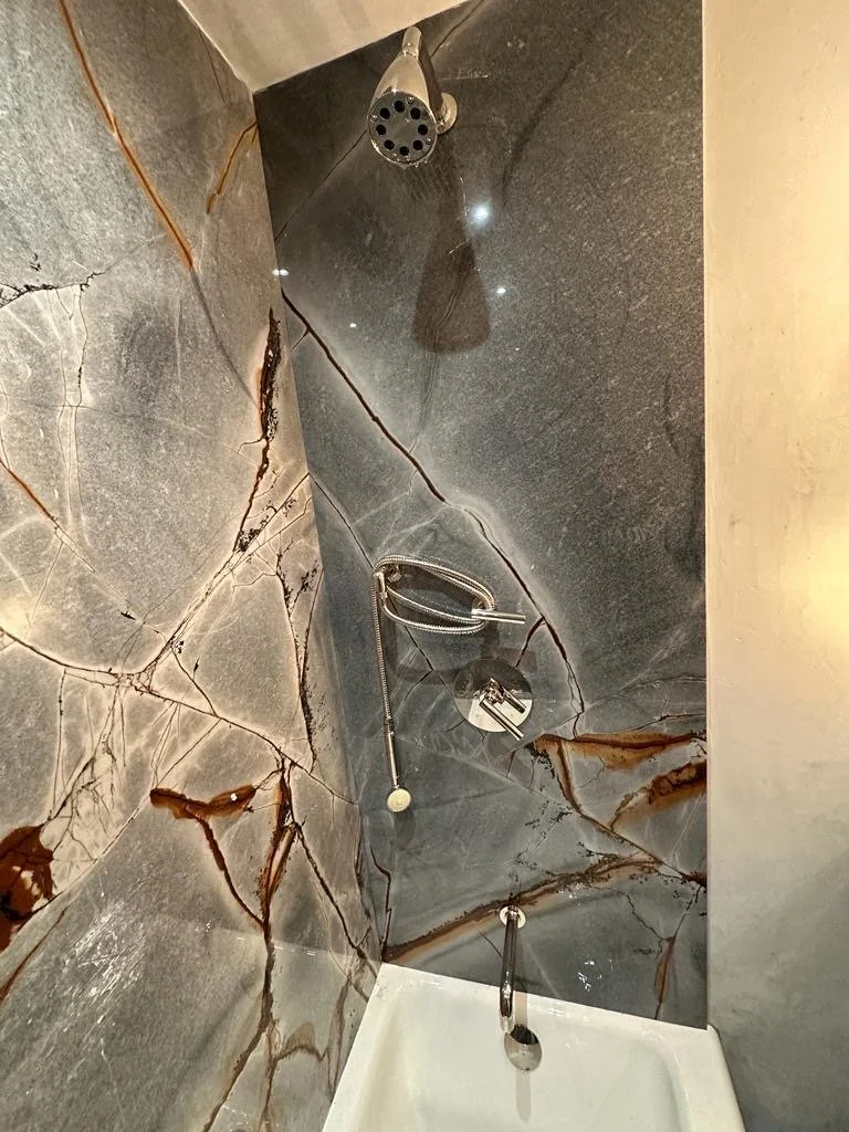 View of a modern shower with dark stone walls featuring reddish-brown veining, a handheld showerhead, a soap dish, and a white bathtub beneath.