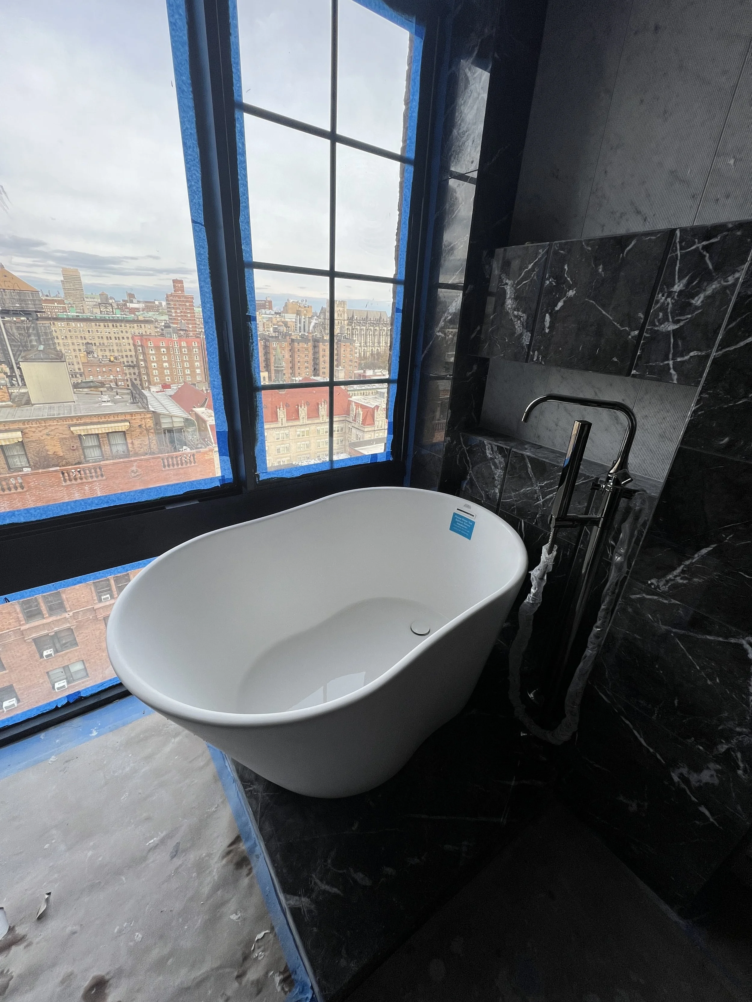 A modern bathroom with a freestanding bathtub near a large window overlooking a city skyline, with black marble tiling behind the tub.