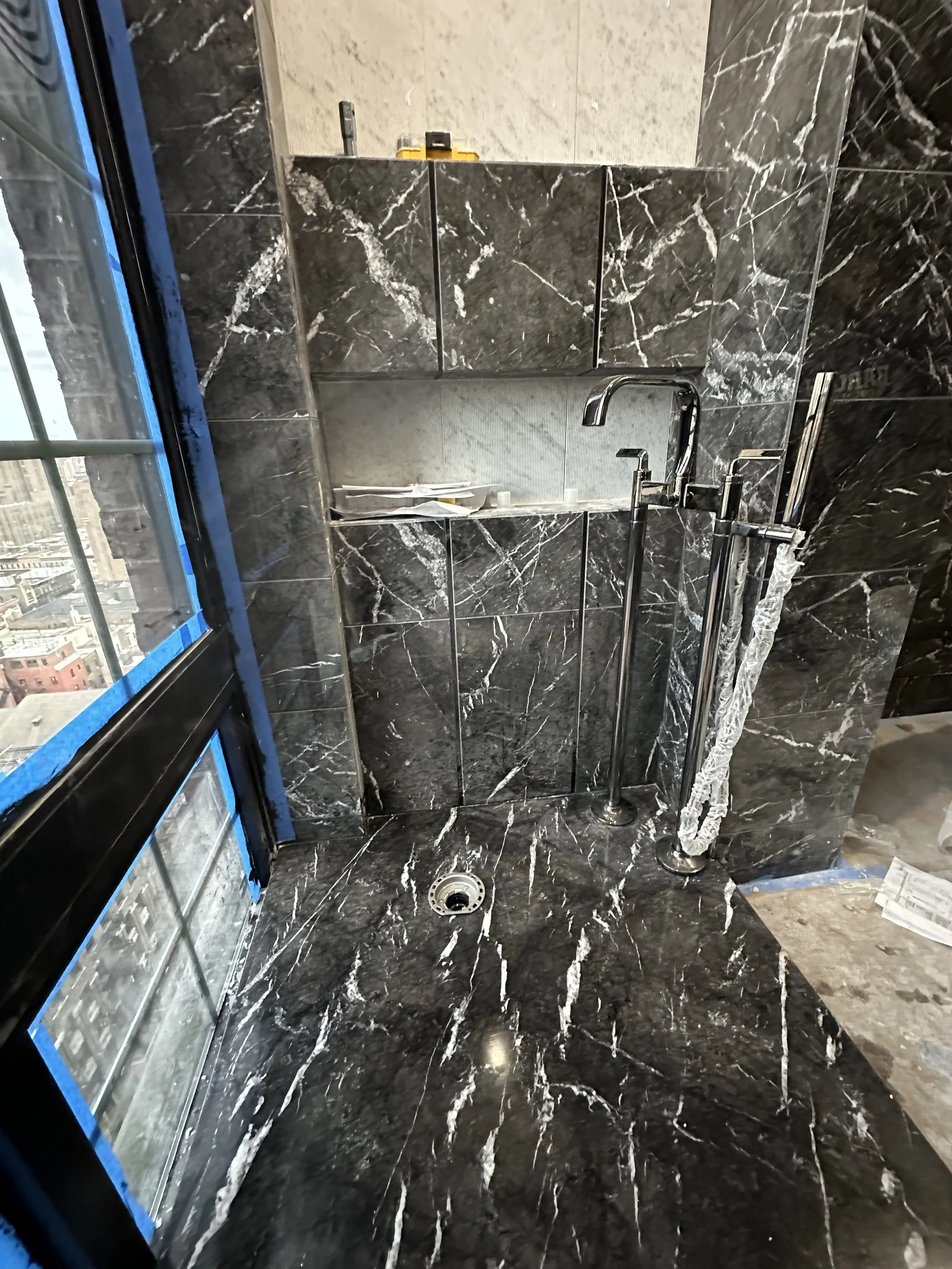 Bathroom shower area with black marble tiles, a wall-mounted small shelf, and two shower fixtures. The image also shows a drain on the floor, a window with blue tape around the frame, and some construction tools on the shelf.