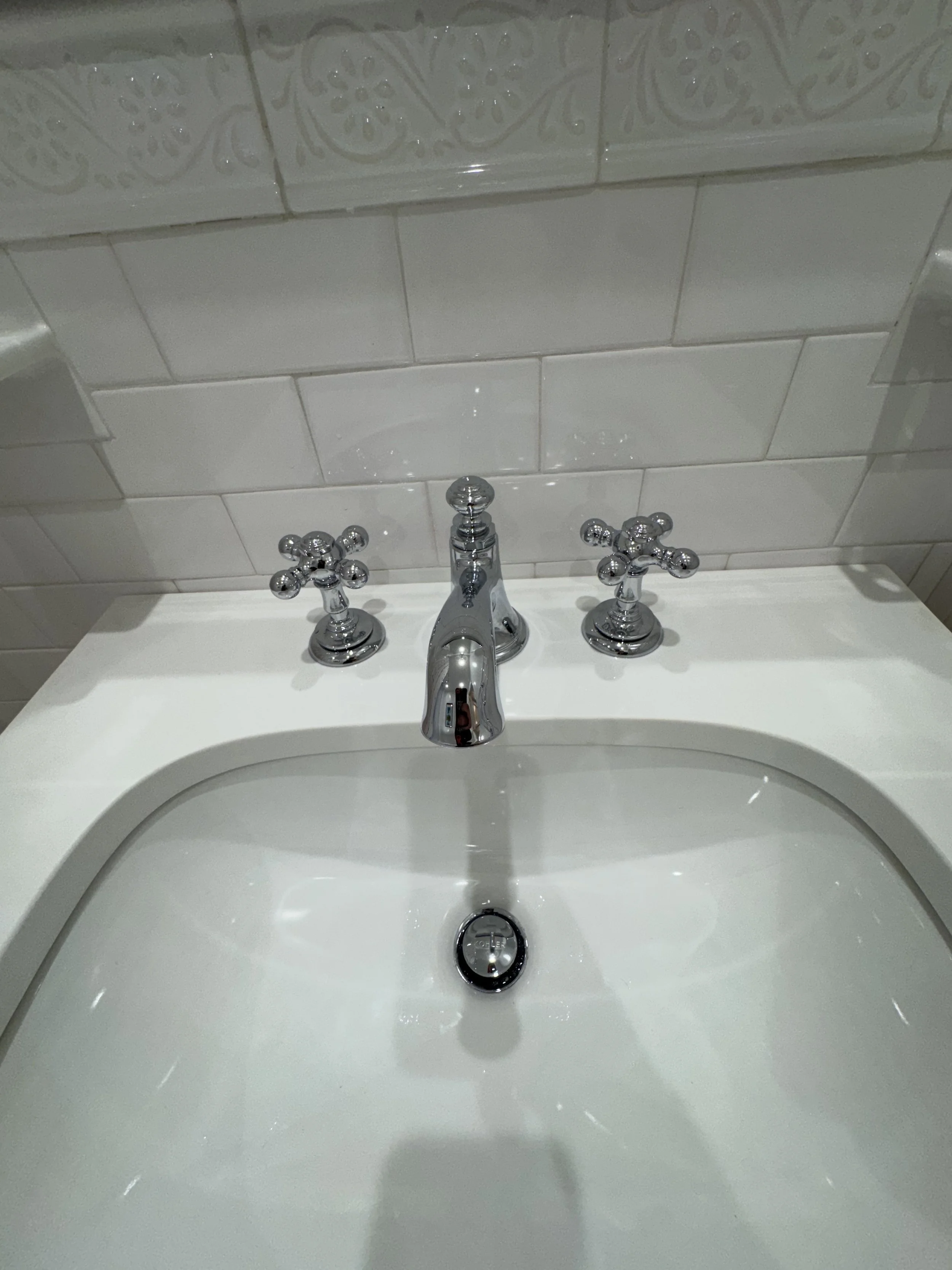 A white bathroom sink with a chrome faucet and two crossed chrome handles on each side. The sink is against a tiled wall with white decorative tiles visible at the top.