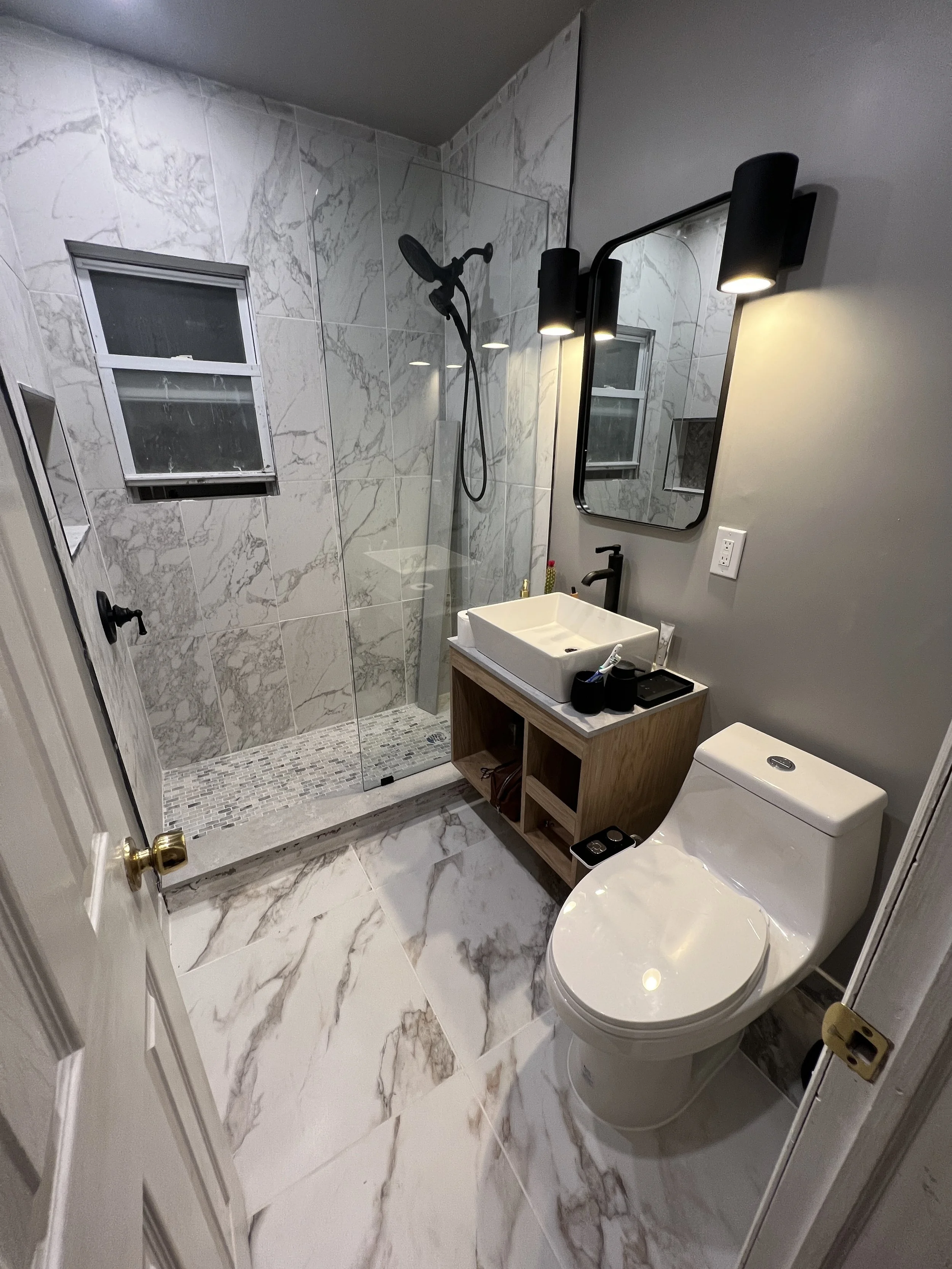 Modern bathroom with marble tiles, glass-enclosed shower, wall-mounted black light fixtures, rectangular sink on wooden vanity, and white toilet.