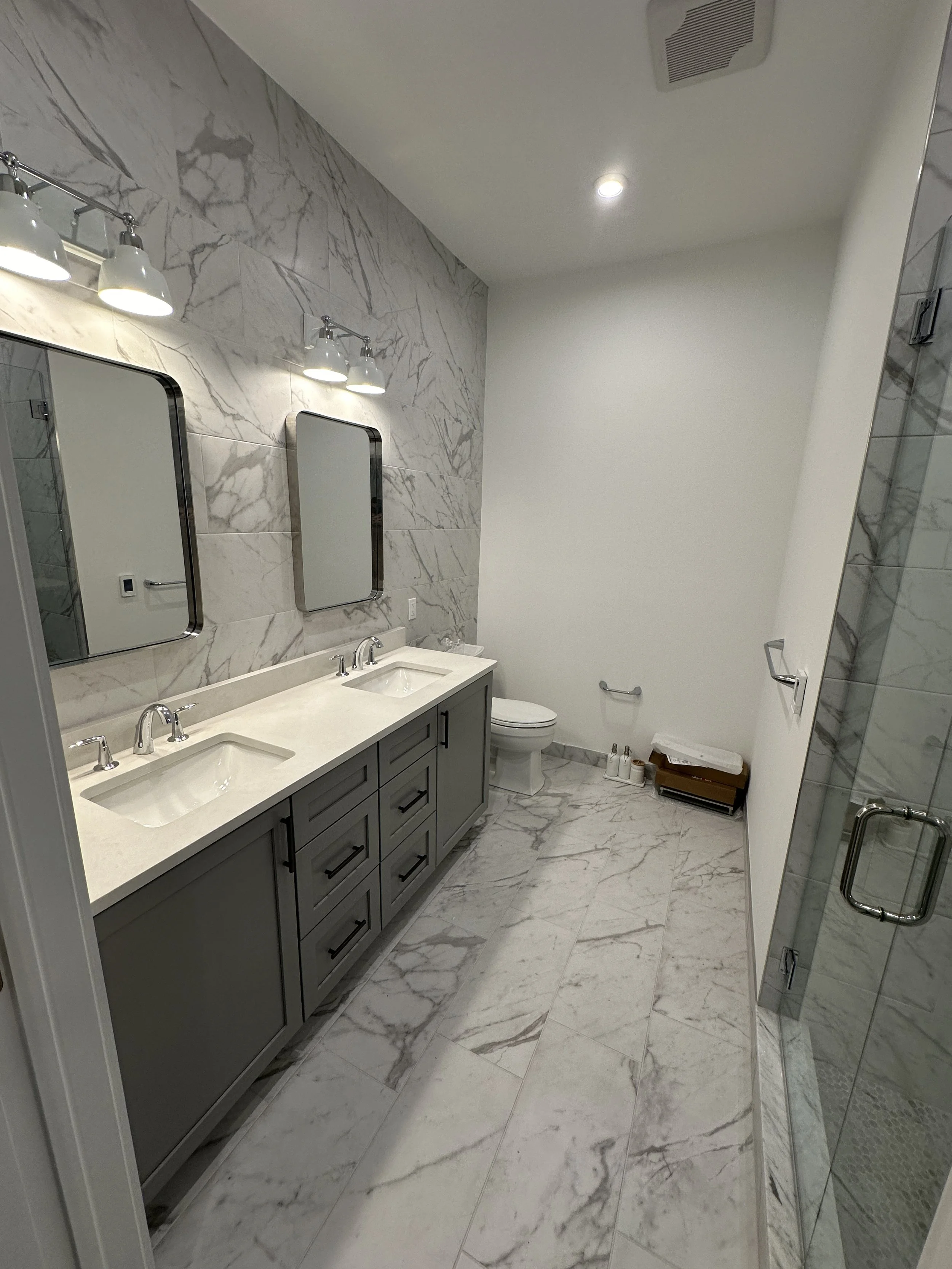 Modern bathroom with marble floor and walls, double sink vanity with gray cabinets, two mirrors, wall-mounted light fixtures, toilet, and glass shower with marble tiles.