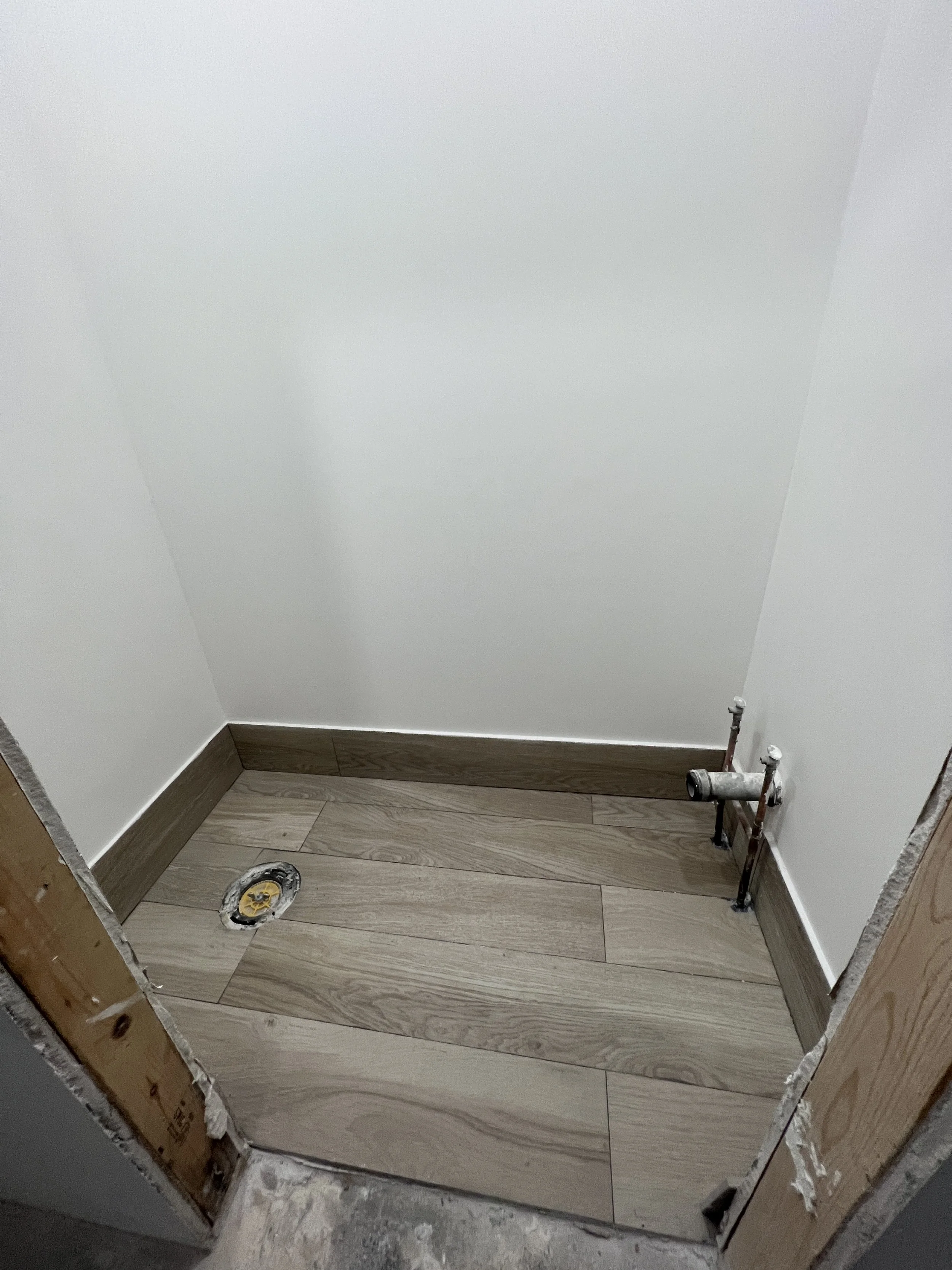 Empty bathroom corner with water pipe installations and a floor drain, walls painted white, baseboard molding, and unfinished door frame.